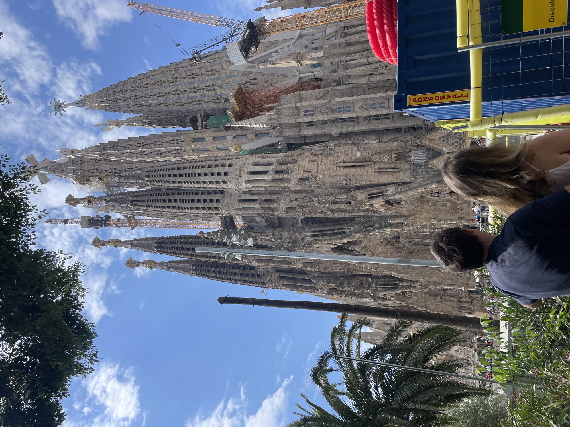 This media file captures a sunny daytime view of the Sagrada Familia basilica in Barcelona, Spain, seen from a ground-level perspective. The grand, intricate stone architecture of the basilica dominates the background, with multiple spires reaching towards a clear blue sky dotted with scattered white clouds. Several large construction cranes are visible near the upper sections of the basilica, indicating ongoing work on the famous landmark.
In the foreground, partially obscured by green foliage, are two people, seen from behind, appearing to be observing the basilica. One person with blonde hair is positioned to the right, and another with dark hair is further right. Trees and palm fronds frame the bottom and left edges of the image, adding a natural element to the urban setting.
A blue container-like structure with yellow accents and red corrugated tubing is visible on the right side of the image, likely a temporary construction facility. Text visible on this blue container reads "PONS DE VALL" and "LEGOTTI." Another yellow sign attached to it displays the word "Disculpa." The bright lighting suggests it is midday or early afternoon. The overall scene depicts a vibrant tourist or visitor experience at an iconic, still-developing architectural marvel.