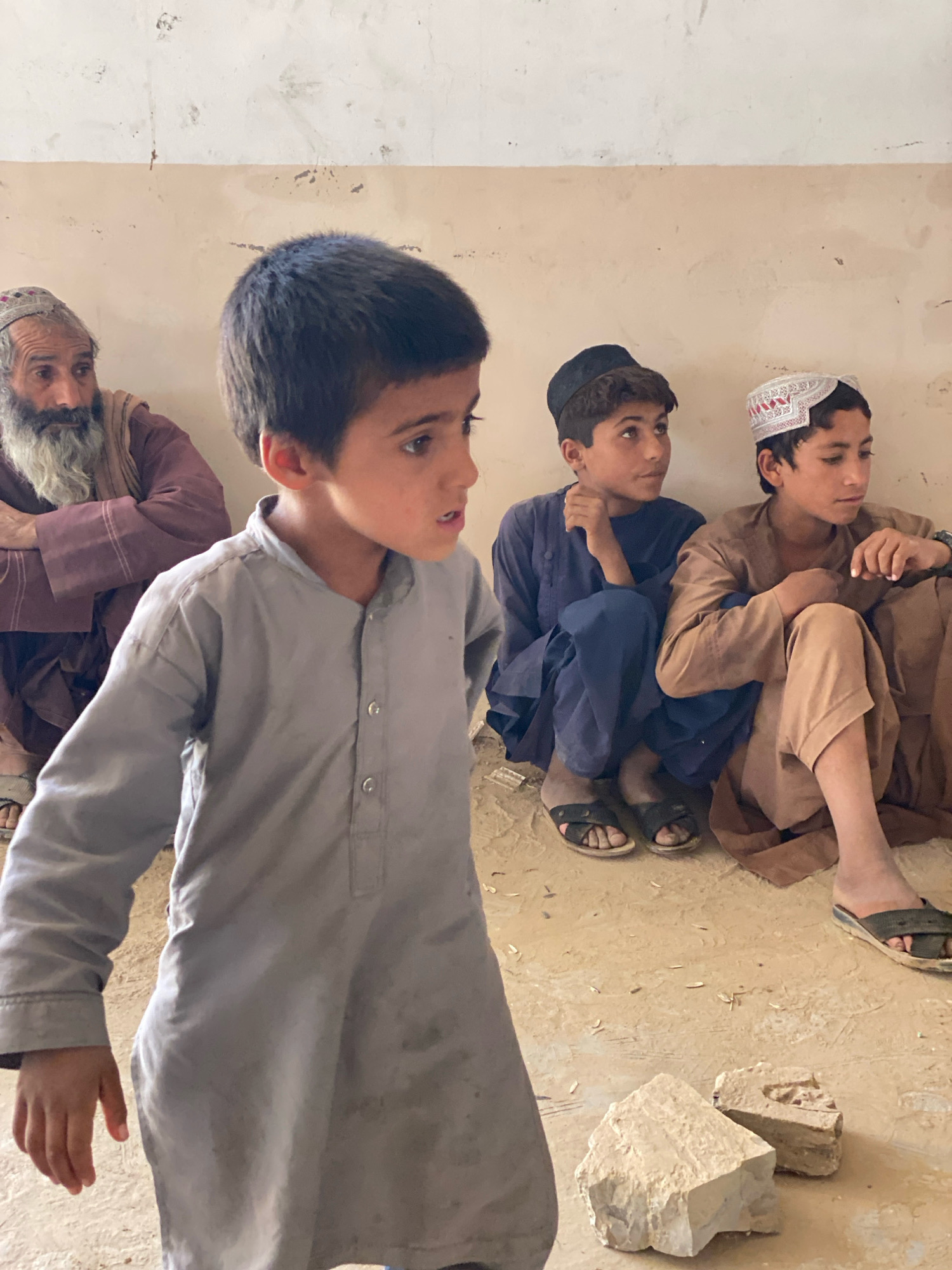 In Pasab, Afghanistan, an indoor scene depicts four males of varying ages. In the foreground, a young boy in a grey tunic faces left, his mouth slightly open. Behind him, an older man with a long grey beard and a patterned skullcap sits to the left, wearing a reddish-brown garment. To the right, two boys are seated on the floor. The boy on the left wears a dark blue tunic and a black cap, while the boy on the right is in a light brown tunic and a white and red patterned skullcap. The environment appears to be a room with light-colored, textured walls and a dirt or concrete floor. Several small, light-colored stones or debris are scattered on the floor in the foreground and middle ground. The lighting is consistent with daylight, casting soft shadows.