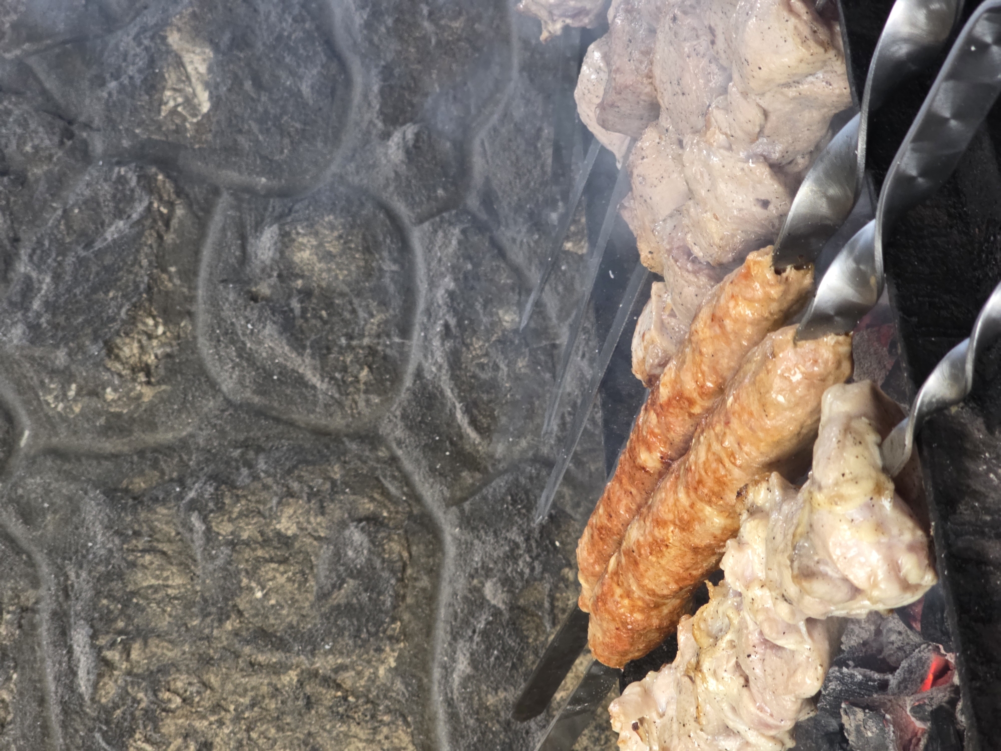 This media file captures a close-up, overhead view of food being grilled on skewers over hot coals. The foreground is dominated by the cooking meat, with several skewers visible. One skewer holds what appears to be sausages, which are golden brown and slightly glistening. Adjacent to the sausages are skewers with chunks of raw, seasoned meat, likely pork or chicken, with visible black pepper. A few pieces of the raw meat are already starting to brown on the edges from the heat. Wisps of smoke rise from the grill, obscuring parts of the scene and adding a dynamic element to the image.
The background features a rough, textured stone wall, likely a barbecue pit or outdoor kitchen structure. The stones are varied in color, ranging from dark gray to lighter shades of gray and tan, with a natural, uneven surface. The lighting suggests it is daytime, with natural light illuminating the food and the stone. There are no people or animals visible in the frame, focusing solely on the food preparation. The overall impression is one of a home-cooked meal or a casual outdoor gathering in Pidhorodne, Ukraine, centered around a traditional barbecue. The focus on the food and the grilling process evokes a sense of warmth and the anticipation of a delicious meal. No visible text is present in the image.
