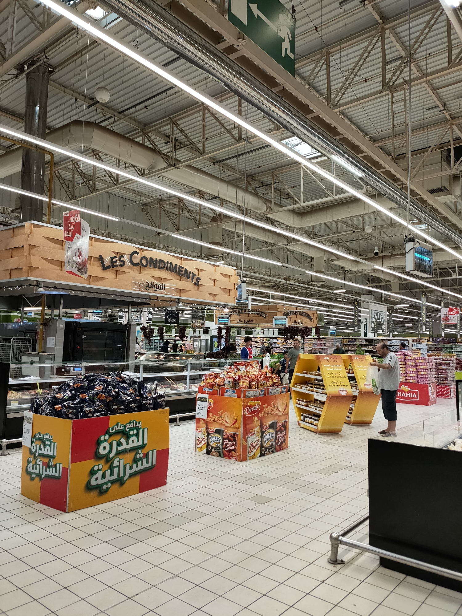 The image captures an indoor view of a brightly lit supermarket aisle, identified by signage as located in Cebalat, Tunisia. The ceiling is a complex network of metal beams and supports, with numerous fluorescent light fixtures illuminating the scene. Prominently displayed above a food counter is a large wooden sign with "LES CONDIMENTS" written in English and Arabic script. In the foreground, a large yellow and red display bin is filled with bags of Doritos chips, with Arabic text on the bin. Further into the aisle, various food products are on display, including snacks, jars of honey, and packaged goods. Several shoppers are visible, some browsing products and others walking through the aisle. A man in a grey t-shirt and dark shorts stands near a display of honey jars, holding a white plastic bag. The floor is tiled in a white grid pattern. The overall impression is of a busy, well-stocked retail environment.