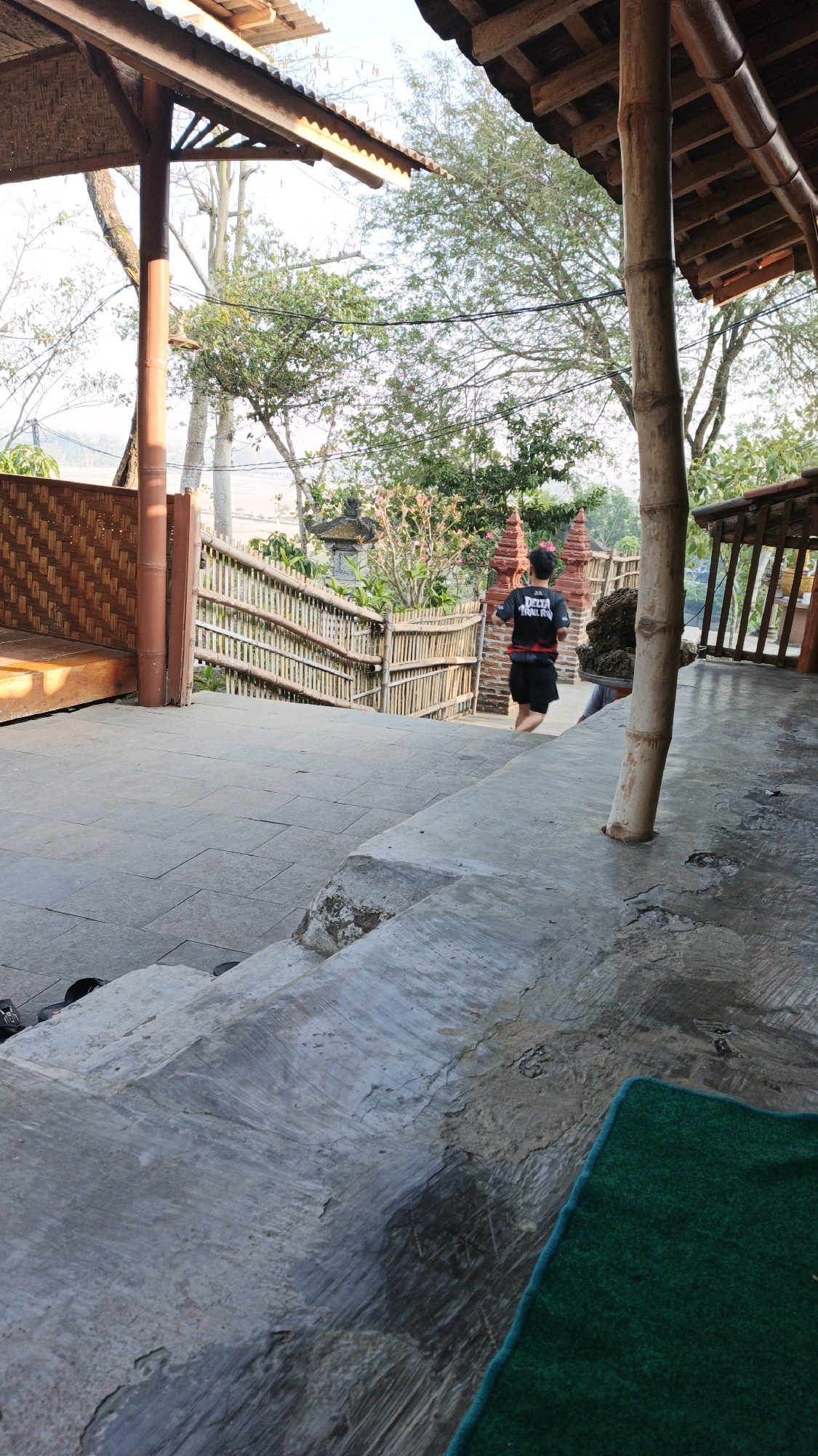 The image depicts an outdoor scene at a location that appears to be in Pojok, Indonesia. In the foreground, there are concrete stairs, some of which are cracked and worn, with a dark shadow cast across them. To the left of the stairs, a section of a wooden platform with woven bamboo walls is visible. In the middle ground, a young person with dark hair, wearing a dark t-shirt with white lettering and black shorts, is seen running up a set of concrete stairs. They are in motion, with their right leg extended forward. Behind the runner, a bamboo fence lines a pathway that leads upwards, with trees and foliage visible beyond the fence. Two small red structures, possibly decorative pillars, stand near the fence. The background features more trees and vegetation under a bright sky, suggesting it is daytime. Wooden structures with thatched roofs are also present in the upper portion of the image, framing the scene from above. The overall impression is that of an outdoor walkway or path in a natural setting, possibly a tourist site or residential area.