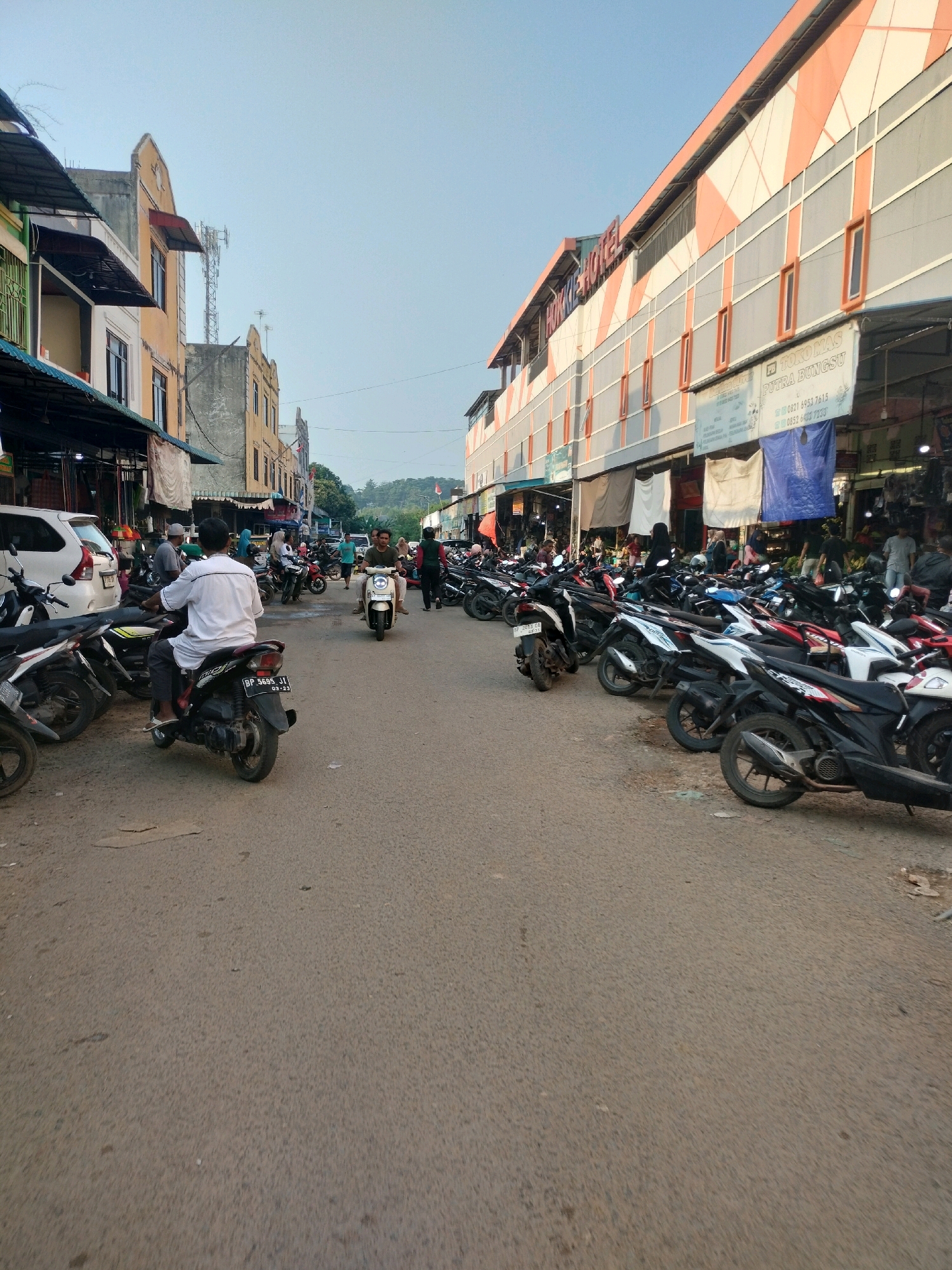 The image depicts a street scene in Batam, Indonesia, characterized by a mix of commercial buildings and parked motorcycles. On the right side of the street, a multi-story building with an orange and grey facade features signs for "HONKIE HOTEL" and "TOKO MAS PUTRA BUNGSU". The ground floor of this building appears to be a market or shop area with various stalls and blue tarpaulin coverings. A large number of motorcycles are parked along the right side of the street, forming a dense row.
On the left side of the street, there are buildings with awnings and what appears to be a market or shop fronts. A white car is parked on the far left. Several people are present in the scene. A man wearing a white shirt is riding a black motorcycle in the foreground, facing away from the camera. Another person is riding a white scooter in the middle distance. Other individuals are visible walking or standing near the shops and motorcycles. The street itself is paved and appears to be in moderate condition, with some patches and debris. The sky is clear and pale blue, suggesting it is daytime, likely late afternoon or early morning given the lighting. The overall impression is that of a busy commercial area with a strong presence of two-wheeled transportation.