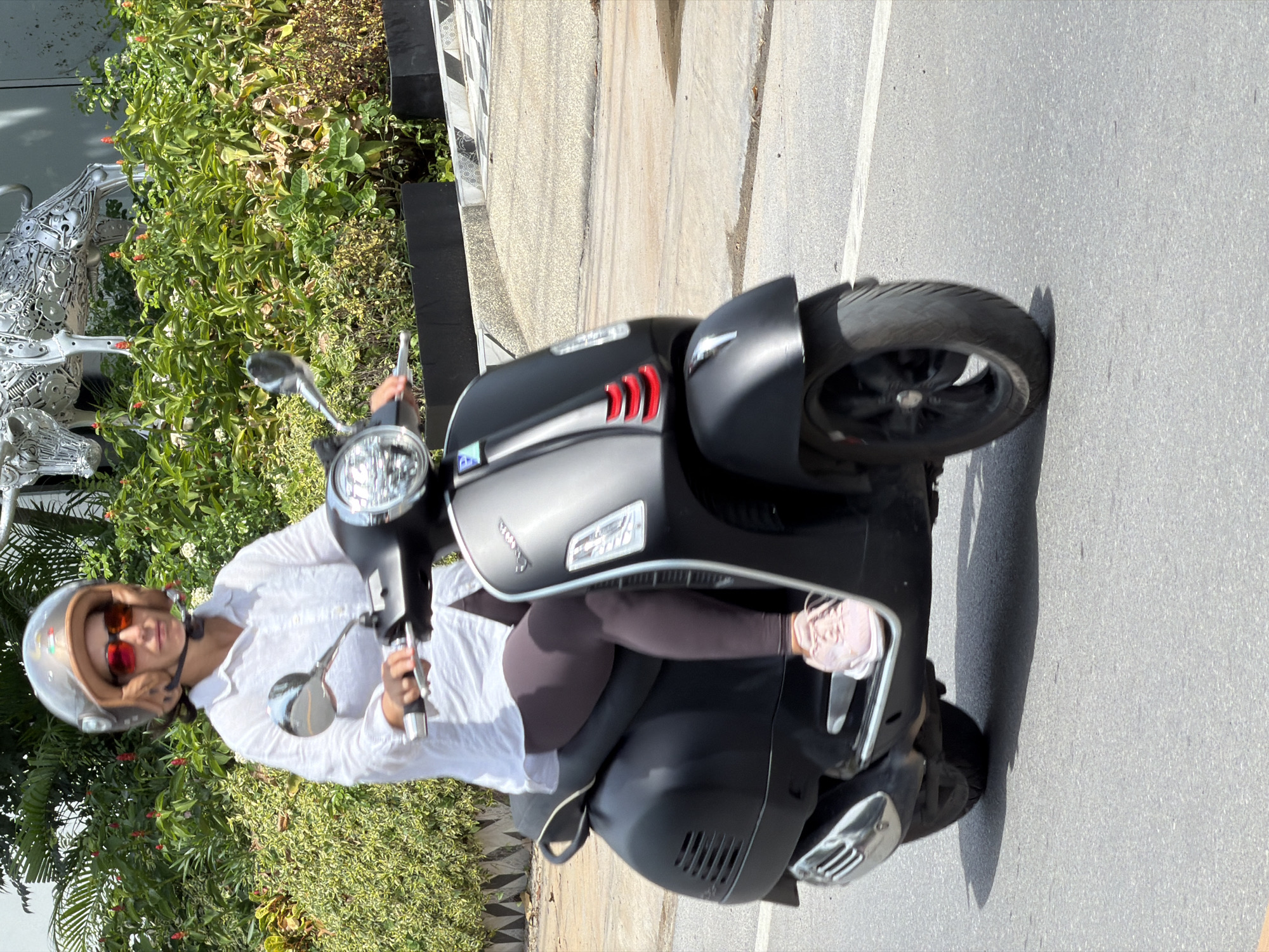 A person wearing a white long-sleeved shirt, dark leggings, pink sneakers, sunglasses, and a silver helmet is riding a matte black Vespa scooter. The scooter has red accents on the front. The person is holding the handlebars and appears to be looking forward. The background shows lush green bushes and trees, with a glimpse of a metallic sculpture of a bull in the upper left corner, and a building with a patterned facade behind the foliage. The scene is set on a paved road in Baan Bo Phut, Thailand.