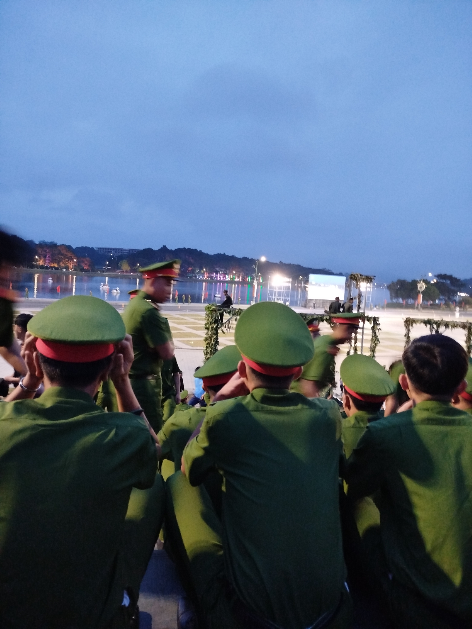 The image depicts an outdoor gathering of individuals, likely military or police personnel, wearing green uniforms and distinctive green caps with red bands. The perspective is from behind a group of these individuals, looking towards a body of water with a cityscape in the background. The sky is overcast and twilight, with artificial lights visible in the distance along the shoreline and on buildings. The setting appears to be an open area, possibly a plaza or park, with some decorative elements like vines on scaffolding visible. The overall atmosphere suggests an event or ceremony taking place in an urban environment, specifically Da Lat, Vietnam, given the context provided. The presence of a large body of water and the surrounding hills are characteristic of the city.