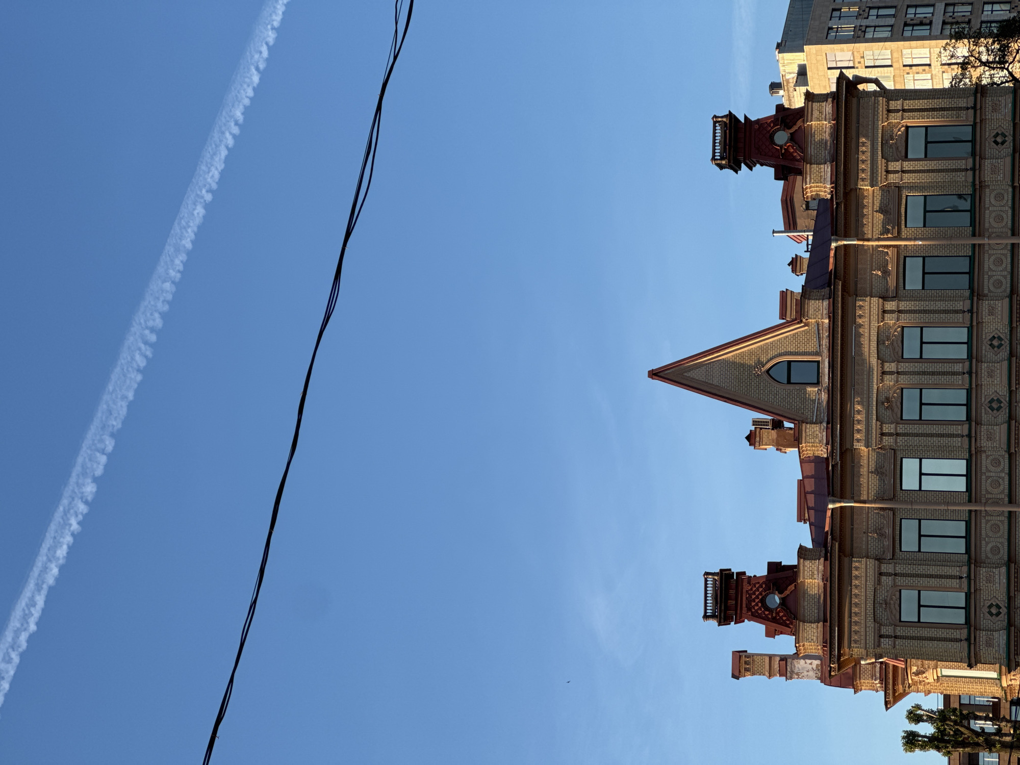 The image depicts the facade of an ornate yellow brick building with a red roof and decorative elements, likely in Saint Petersburg, Russia. The building features multiple chimneys and an elaborate central turret. To the right, a modern concrete building with numerous windows is visible. The sky is a clear blue with a faint contrail from an airplane and a single bird in the distance. Overhead, two black electrical wires stretch across the frame. The scene is illuminated by natural light, suggesting it was captured during the day, possibly late afternoon or early morning due to the warm light on the building.