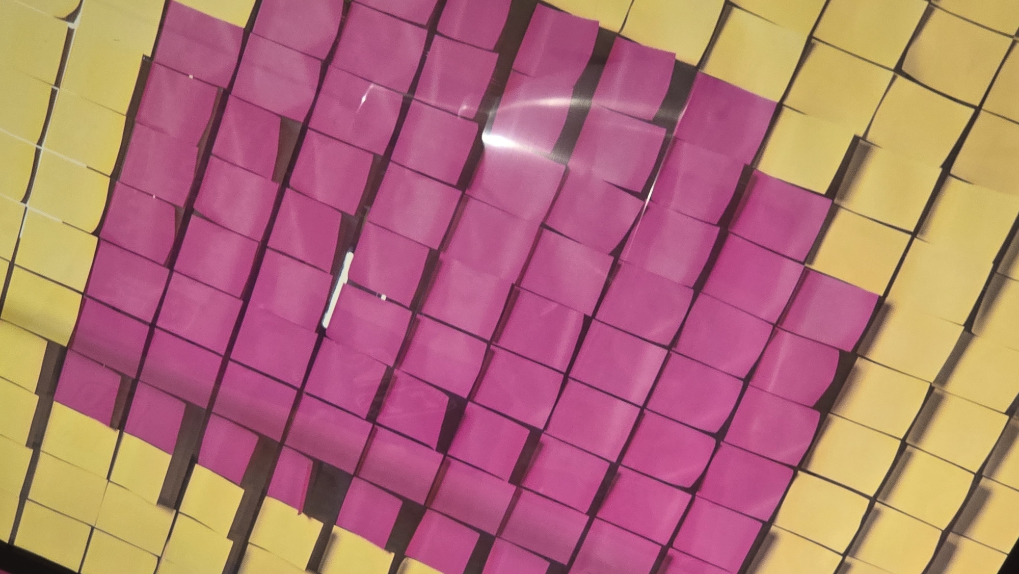 The image shows a wall covered with yellow and pink square sticky notes arranged in a pattern. The sticky notes form a large, pixelated shape resembling a pink diamond or heart on a yellow background. The arrangement of the sticky notes creates a textured, three-dimensional effect, with some notes casting shadows. The lighting in the image is bright, causing some glare on the surface of the sticky notes, particularly in the center of the pink area. The perspective is from a high angle looking down at the wall. There are no people visible in the image. The location of this artwork in Chaikovsky, Russia, is not discernible from the image itself.