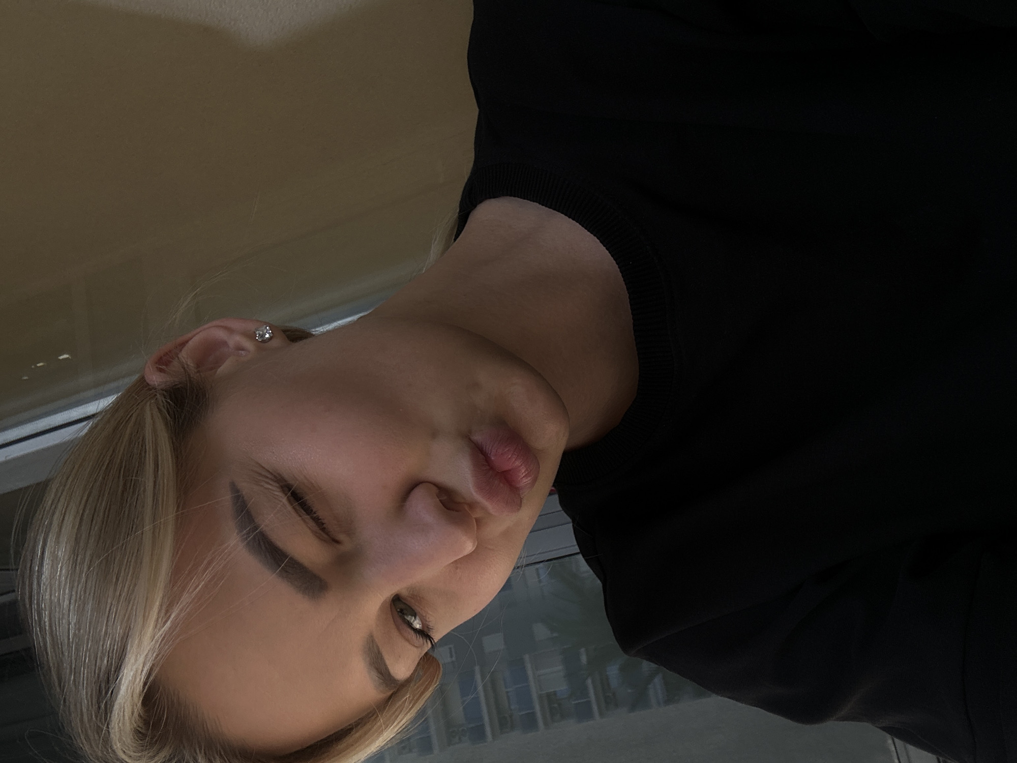 A close-up portrait shows a person, likely female, with fair skin and blonde hair, wearing a black long-sleeved top or sweater. The image is rotated 90 degrees counter-clockwise, making the subject's head appear significantly tilted. She is looking downwards and to her left, with her lips slightly pursed in a pout or contemplative expression. Her visible eye, partially open, reveals a green iris and distinct eyelashes. She has dark, well-defined eyebrows. A small, sparkling stud earring is visible in her left earlobe. The background is softly blurred, showing indistinct indoor elements and reflections of what appear to be outdoor buildings or structures, possibly seen through a window. The lighting suggests an indoor setting, likely during the day. No text is visible in the image. The location context for this media file is Budva, Montenegro.