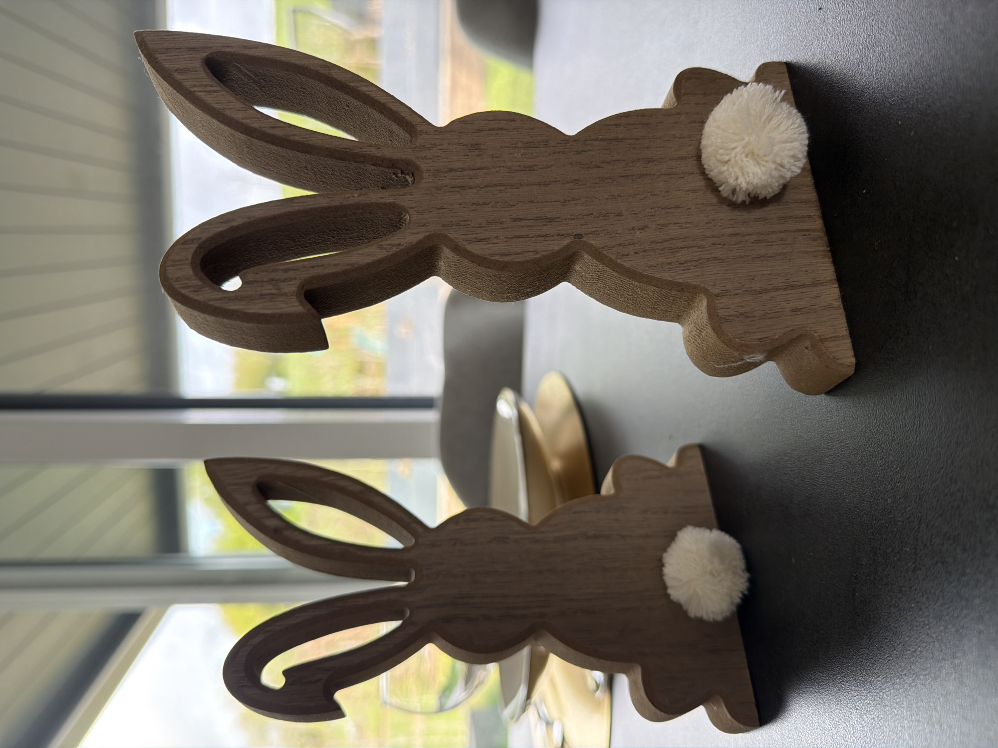 Two wooden rabbit figures with fluffy white tails are sitting on a dark surface. The figures are light brown in color, and the window in the background reflects a natural setting. The location is Junglinster, Luxembourg.