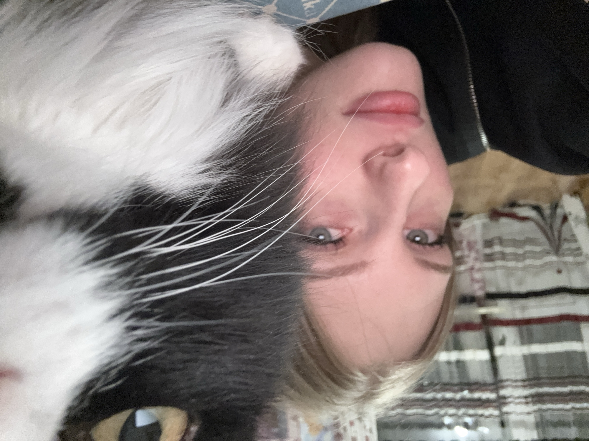 A black and white cat is lying next to a light-skinned woman with blond hair. The image is rotated 180 degrees. The woman is wearing a black zip-up jacket. The location is undetermined.