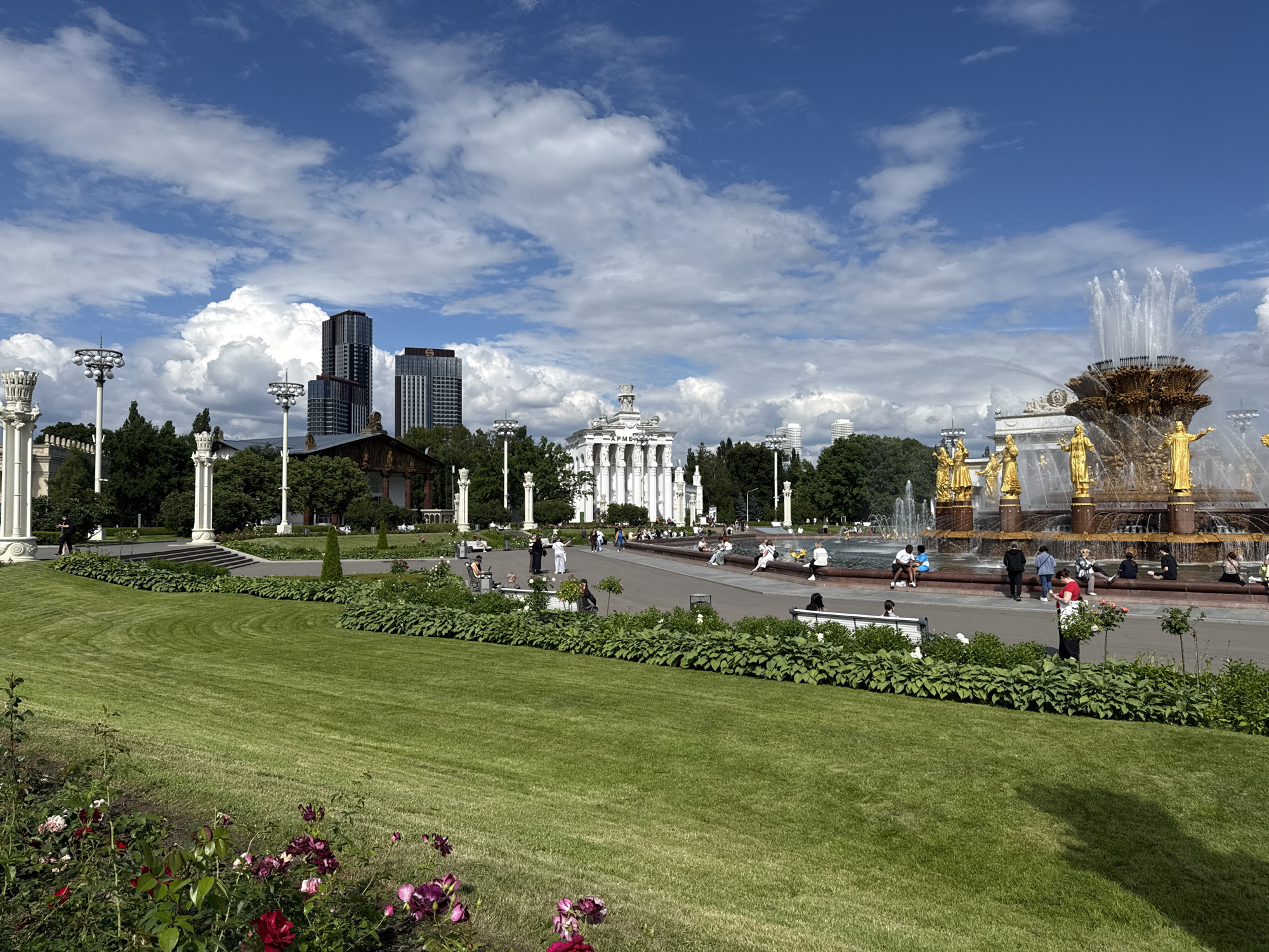 This is a photograph of the Friendship of Nations Fountain at VDNKh in Moscow, Russia. In the foreground, a lush green lawn slopes towards a flowerbed with red and purple roses. Behind the lawn, a pathway is lined with manicured hedges and people are seated on benches. Further back, the iconic Friendship of Nations Fountain is in full operation, with water cascading from its golden structure, adorned with statues of individuals representing Soviet republics. To the left of the fountain, a grand white pavilion with columns and statues is visible. In the background, modern high-rise buildings rise above the trees, contrasting with the neoclassical architecture of the exhibition grounds. The sky is blue with scattered white clouds.