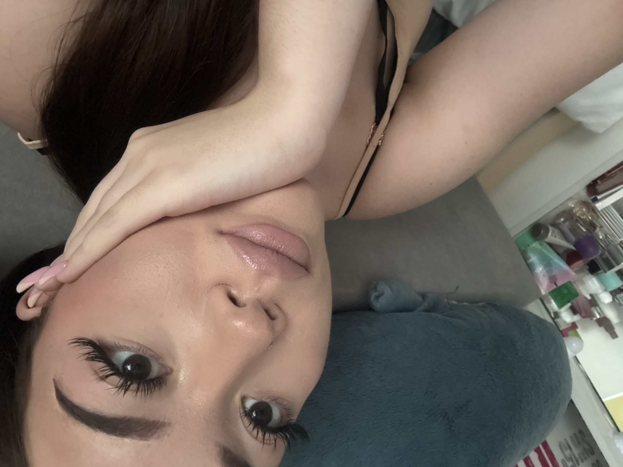 A close-up shot captures a woman with long brown hair, posing with her hand near her face. She is wearing makeup, including noticeable eyelashes and lip gloss. The image is taken at an unusual angle, with the woman's face tilted. Part of her shoulder and a thin black strap, possibly from a garment, are visible. A blue pillow is present in the lower portion of the frame. The background includes a white surface and various small items, suggesting an indoor setting. There is no location information available to include the city of Naberezhnye Chelny, Russia.