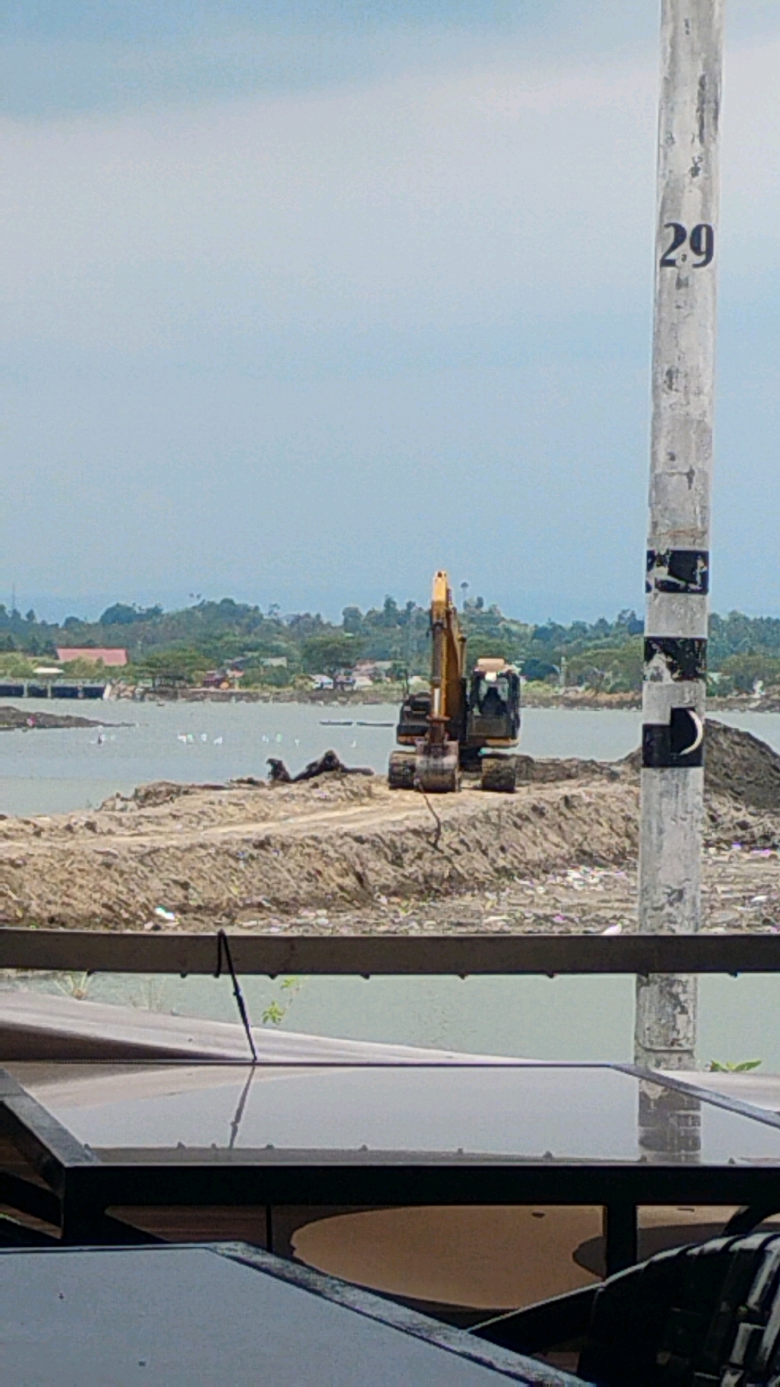 This image, taken in Lhokseumawe, Indonesia, depicts a construction site near a body of water. In the foreground, the edge of a dark, reflective surface, possibly a table or bench, is visible. Beyond this, a light-colored pole with black markings and the number "29" painted on it stands vertically. The background reveals a large body of water with several white birds scattered across its surface. On the far bank of the water, a town with buildings and trees can be seen. A prominent yellow excavator is positioned on a mound of dirt in the middle ground, suggesting ongoing construction or land reclamation work. The sky is overcast.