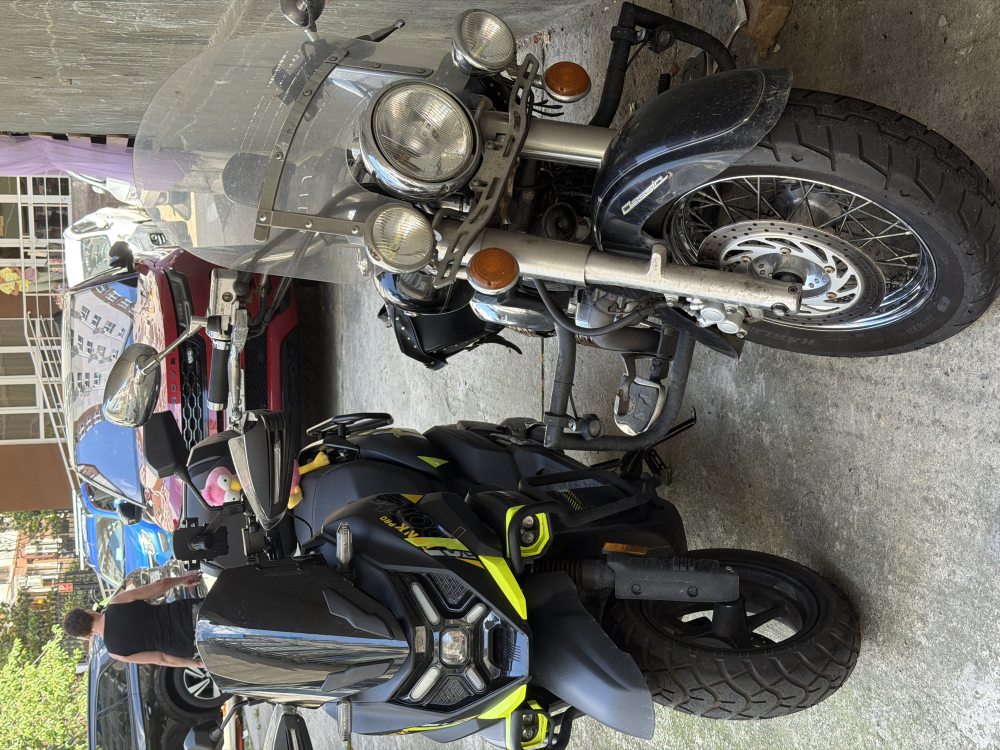 Two motorcycles are parked side-by-side on a concrete surface, possibly in an alley or parking area, in Sochi, Russia. The motorcycle on the left is a modern, black scooter with bright yellow accents and a large, tinted windshield. It features aggressive styling with sharp lines, LED headlights, and knobby tires, suggesting it's designed for varied terrain. A pink flamingo plush toy is visible on its handlebars.
The motorcycle on the right is a classic cruiser with a large chrome windshield, multiple round headlights, and a spoked front wheel. It has a black saddlebag attached to the side. The front tire is thick and black.
In the background, a red SUV is parked, and a blue SUV is visible behind it. A woman wearing a black tank top and shorts is walking away from the viewer, partially obscured by the scooter. Buildings are visible in the distance, indicating an urban or semi-urban environment. The overall impression is one of everyday urban life, with vehicles parked in a typical city setting.
