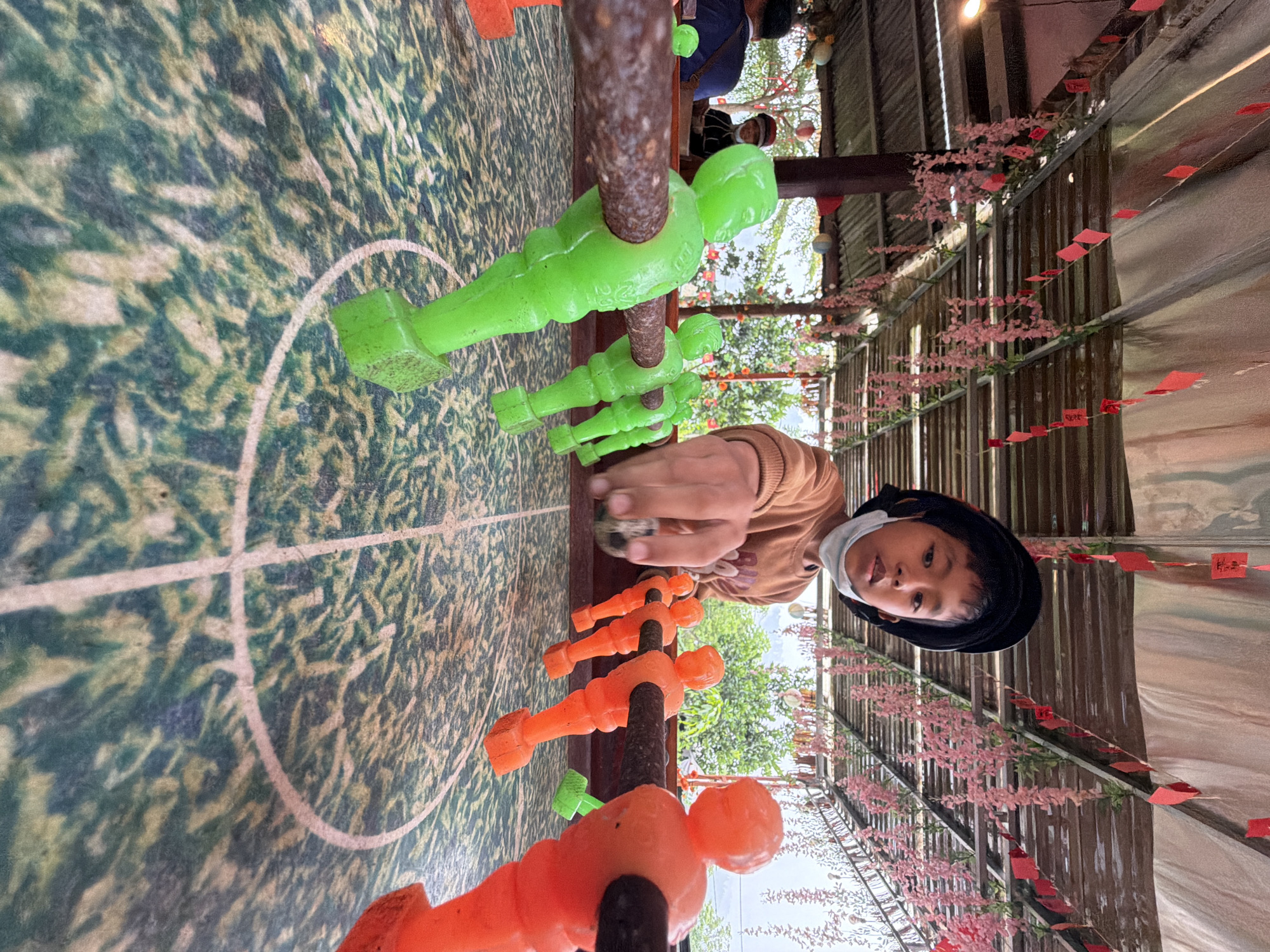 A child wearing a brown sweater, black hat, and a white surgical mask is playing foosball inside a covered outdoor structure. The foosball table features green and orange player figurines. The table has a simulated grass field. The structure is decorated with pink flowering vines and small red hanging tags. The location is likely a tourist attraction in the city of Đà Nẵng, Vietnam. Other people can be seen in the background.