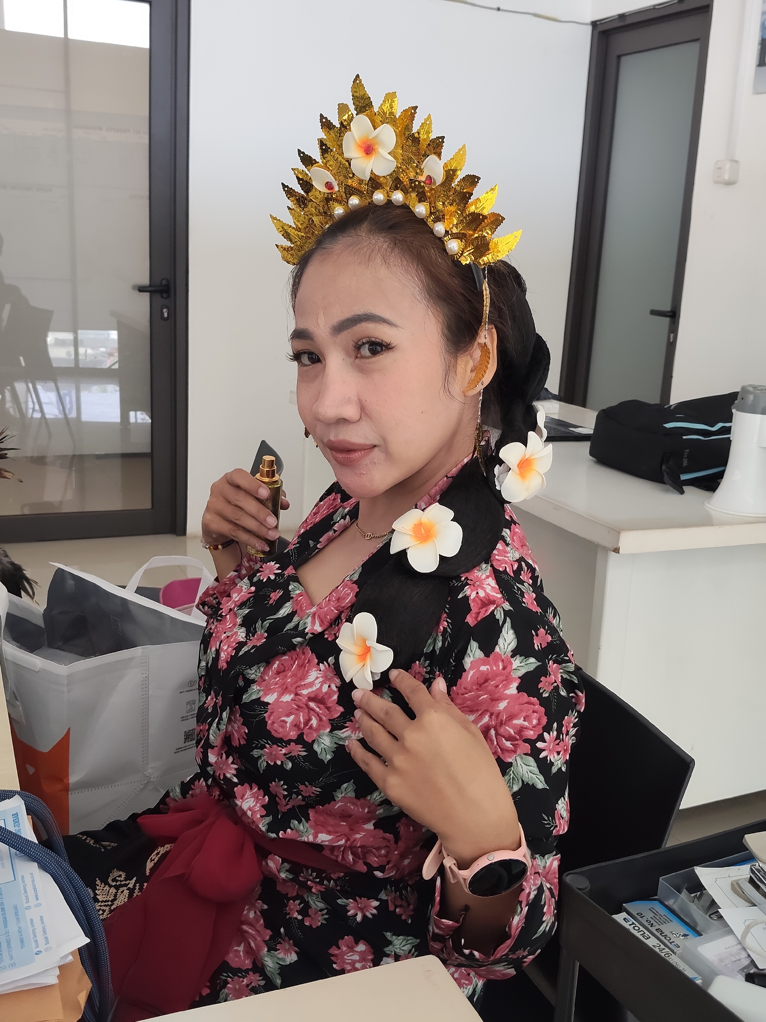 A woman wearing a traditional Balinese headdress and floral patterned dress sits in an office environment. She holds a small bottle of perfume up to her face, looking towards the camera. White flowers are visible in her hair and on her dress. The background includes office furniture, doors, and windows. The scene appears to be in Denasri Wetan, Indonesia.