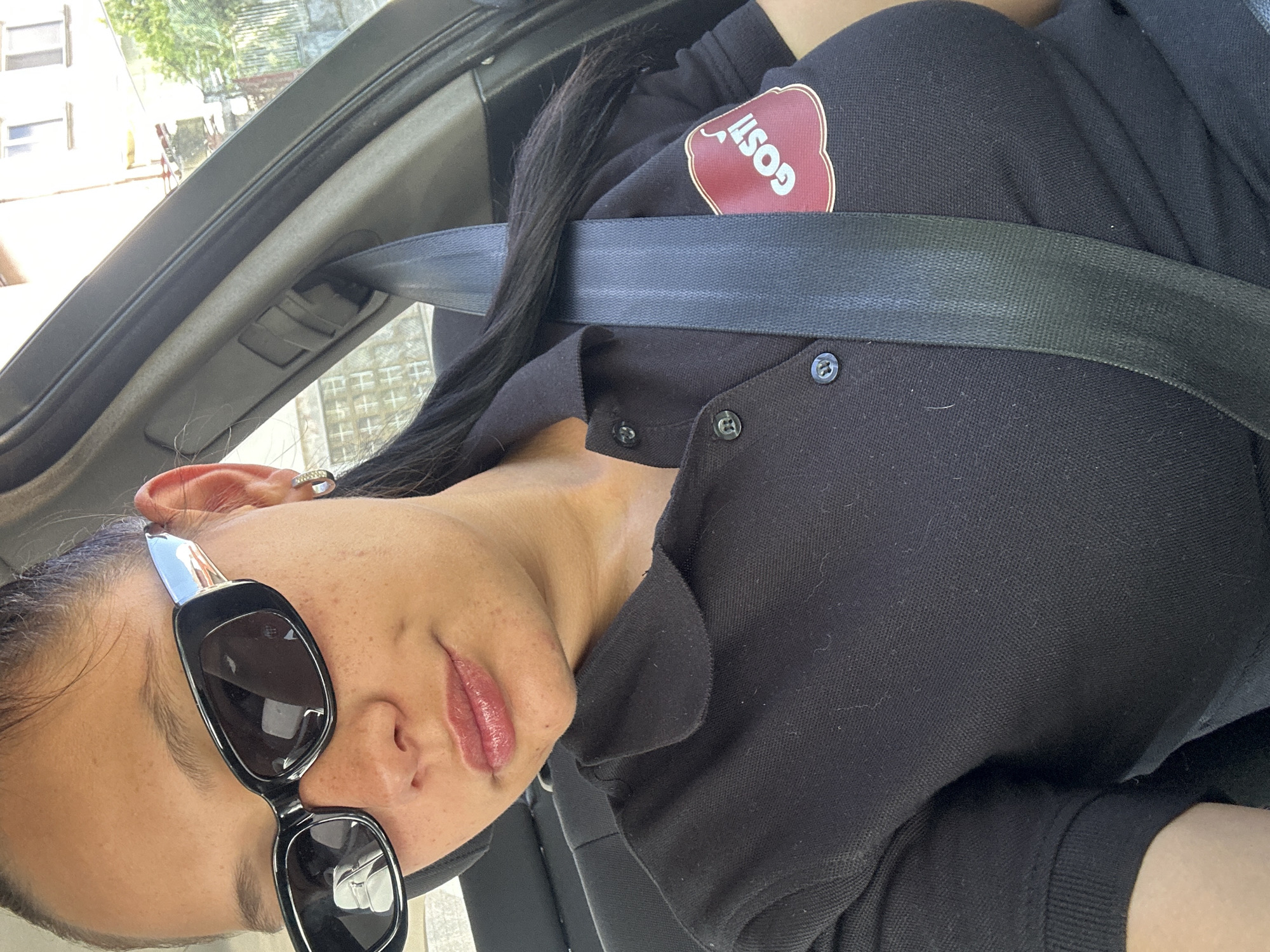 A woman wearing a black shirt with a "Goshi" logo, sunglasses, and a seatbelt is in a car. The location is inferred to be Sofia, Bulgaria. The photo is taken from inside the vehicle.