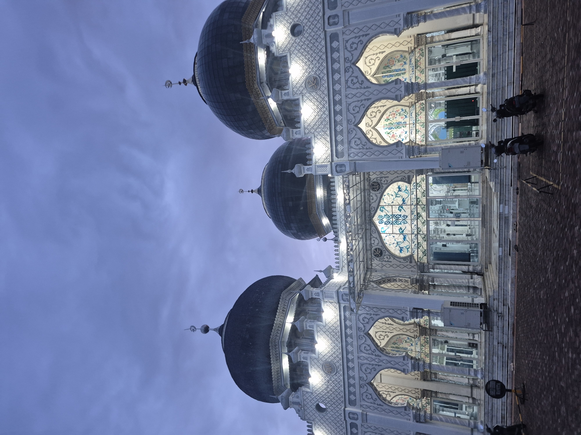 The image shows a mosque with three dark-colored domes and ornate, white architecture against a blue sky, with some lighting. The mosque's facade includes arched windows with decorative patterns. The location is Keudekruenggeukueh, Indonesia. Two parked motorcycles can be seen. The image is rotated.