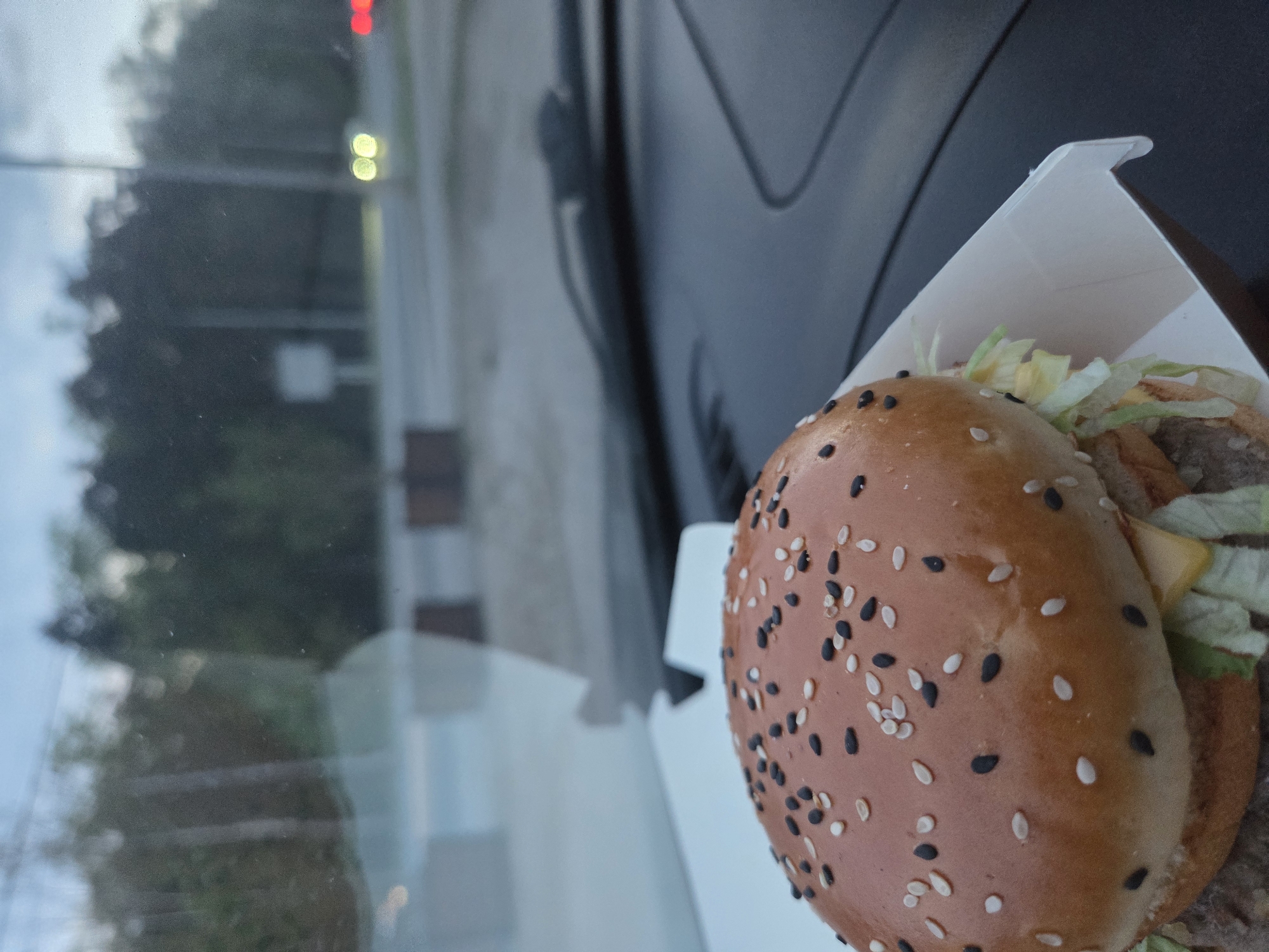 The image shows a burger, likely a Big Mac, in its box, placed on a car dashboard. The burger has a sesame seed bun with both white and black sesame seeds. Lettuce and cheese are visible between the burger patty and bun. Through the car window, an out-of-focus street scene is visible, including streetlights and possibly a traffic light. Based on the appearance of the buildings and street furniture, it's possibly the city of Perm, Russia.