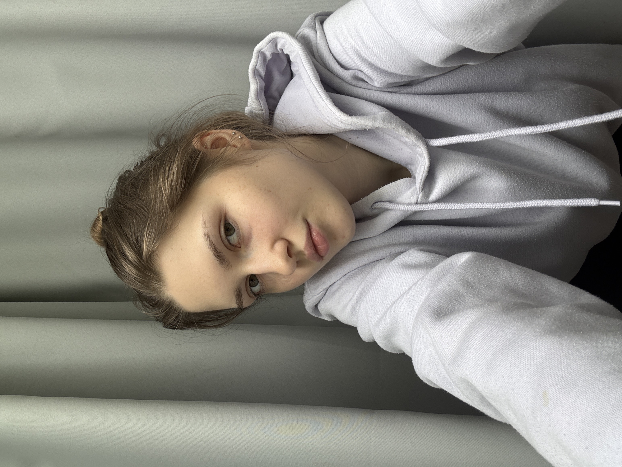 A young woman with fair skin, light brown hair pulled back in a messy bun, and light eyes is wearing a light-colored hooded sweatshirt with white drawstrings. She is looking at the camera with a slight tilt of her head. The background is a neutral-colored, possibly grey, wall or curtain. The image does not contain any information to determine the location in the city of Saint Petersburg, Russia.