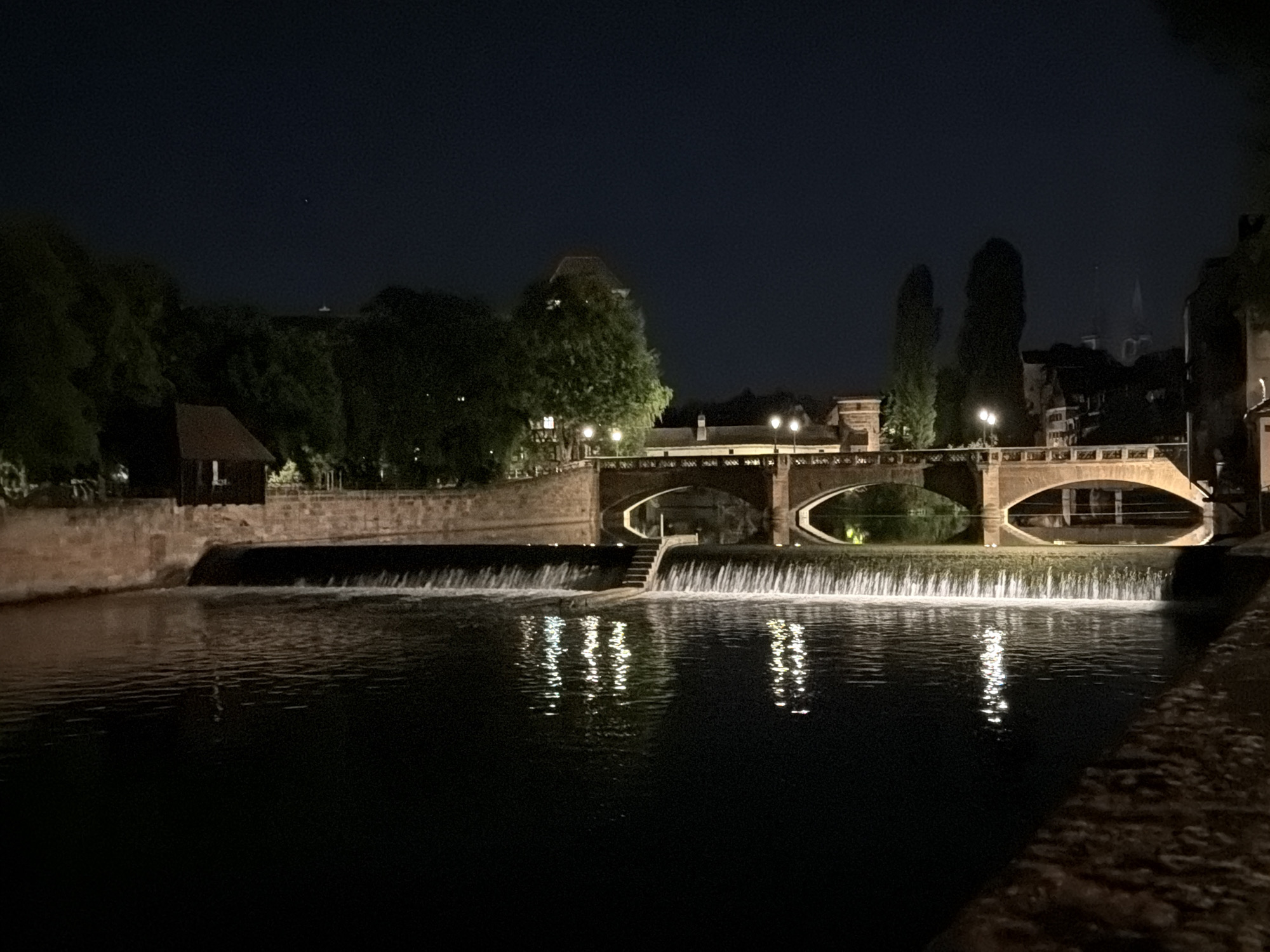 This nighttime photograph captures a serene and picturesque scene along a river, likely the Pegnitz in Nuremberg, Germany, given the context. The sky is dark and clear, with a faint star visible in the top left.
The central feature is a wide river or canal, calm in the foreground but breaking into a dynamic weir or waterfall across the mid-ground. The cascading water is illuminated, appearing white and foamy, and its bright reflections shimmer on the dark water surface below.
Multiple stone bridges with elegant arches span the river beyond the weir. These bridges are lit by numerous streetlights, casting warm glows and creating bright reflections in the water beneath their arches. The architecture of the bridges and the surrounding buildings suggests a historical European setting.
Along the riverbanks, various structures and abundant foliage are visible. On the left, a small, dark-roofed building is nestled amongst trees. More trees line both banks, some illuminated, adding depth and texture to the scene. In the distance, parts of other buildings with traditional pitched roofs can be seen, and a distinct spire or tower is faintly visible against the dark horizon, suggesting a prominent landmark like a church or castle. The overall lighting scheme enhances the architectural details and the natural flow of the water, creating a tranquil and historic atmosphere.
No discernible text is visible in the image.