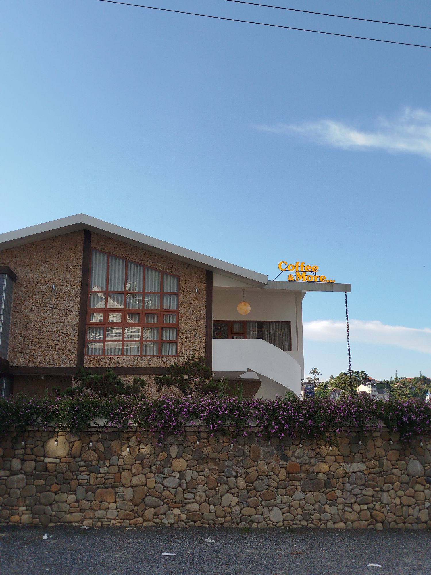 The image shows a modern coffee shop building with a sign that reads "Coffee & More..." in orange lettering against a clear blue sky. The building's exterior is a mix of textured brown stone and white stucco. A large, angular window with wooden framing is prominent on the stone section of the building. In front of the building, a curved stone wall is covered with a dense growth of purple flowers. Below the wall, the ground is covered with dark gravel. The coffee shop is located in Da Lat, Vietnam.