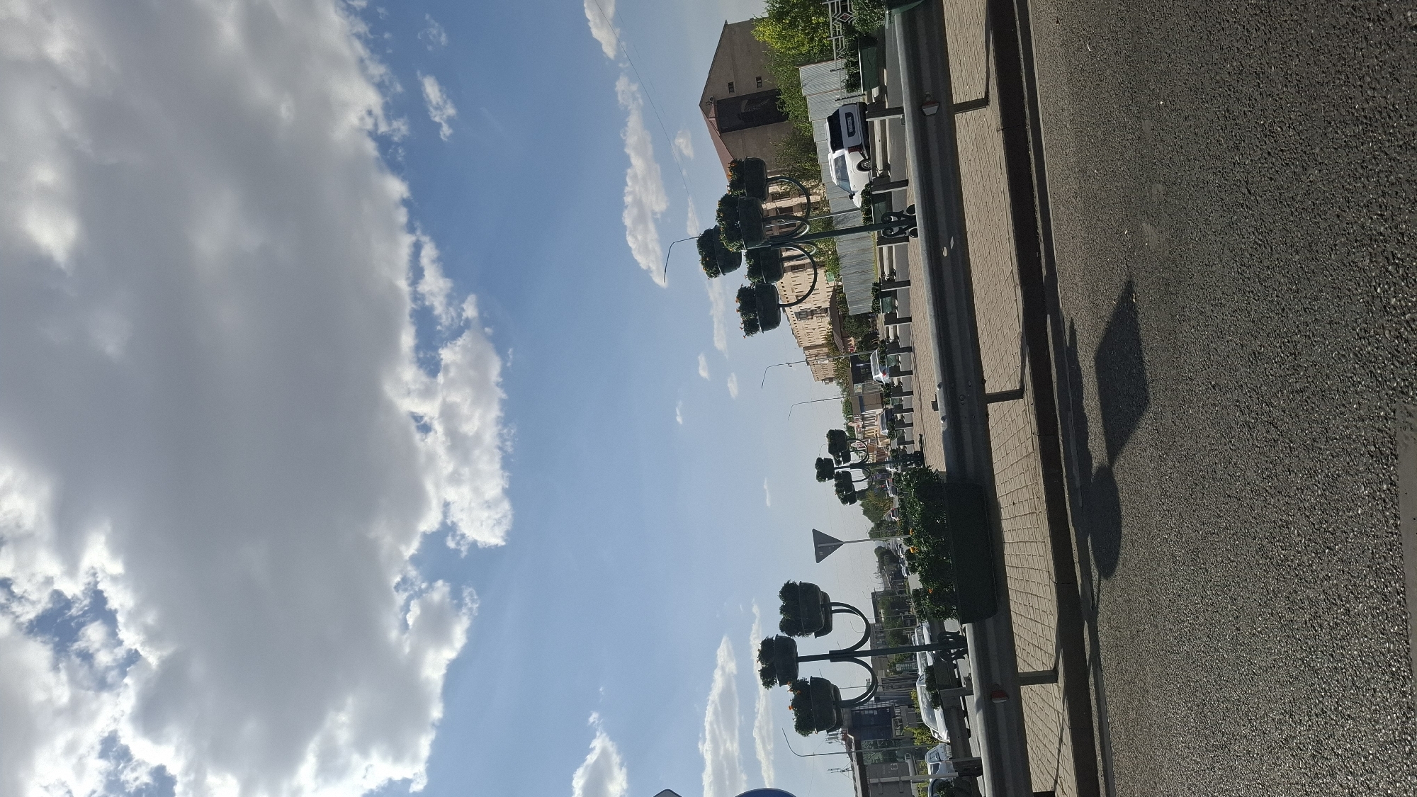 The image shows a street scene in Shymkent, Kazakhstan, on a bright, partly cloudy day. In the foreground, there is a grey asphalt road with a distinct shadow cast upon it. Beyond the road is a paved sidewalk bordered by a metal guardrail. Along the sidewalk, decorative streetlights with potted plants are arranged, with some of the plants in bloom. Further back, there is a row of cars, including a white SUV. Buildings, some with apartment-style facades, and trees line the opposite side of the street. A tall building with a light brown exterior is visible in the middle ground. The sky is blue with scattered white and grey clouds. The overall impression is of a sunny day in a developed urban area.