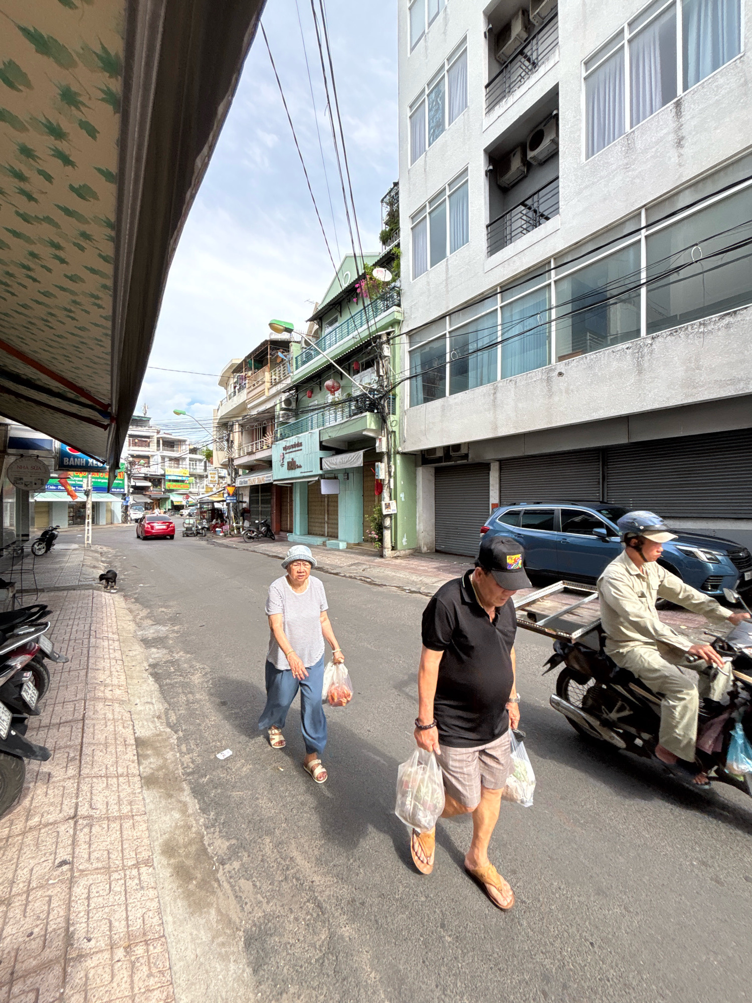 This image captures a street scene in Nha Trang, Vietnam, likely in a commercial or residential area. The perspective is from street level, looking down a moderately wide road lined with buildings and shops. The sky is partly cloudy.
On the right side of the street, a modern, multi-story building with a light gray facade dominates the background. It has numerous windows and balconies with railings. Below the windows, there are large, dark roller shutters, indicating commercial establishments. A blue SUV is parked in front of one of these shutters.
Closer to the foreground on the right, a man wearing a black t-shirt, plaid shorts, a blue baseball cap, and flip-flops is walking towards the viewer. He is carrying two plastic bags filled with groceries. To his right and slightly behind him, a person on a motorcycle, wearing a light-colored shirt and pants and a helmet, is also moving away from the camera. The motorcycle appears to be carrying some cargo in the back.
On the left side of the street, a woman in a striped shirt, blue pants, and sandals is walking away from the camera. She is also carrying a plastic bag of groceries. Adjacent to the sidewalk on the left, there is a red car parked. Further down the street, other vehicles, including motorcycles and scooters, are visible.
The sidewalk on the left is paved with light-colored rectangular tiles. A black dog is visible on the sidewalk. The buildings along the left side of the street are varied in height and style, with awnings and balconies. Some of the shops have signage with Vietnamese text. The overall impression is of a typical day on a busy street in a Vietnamese city.