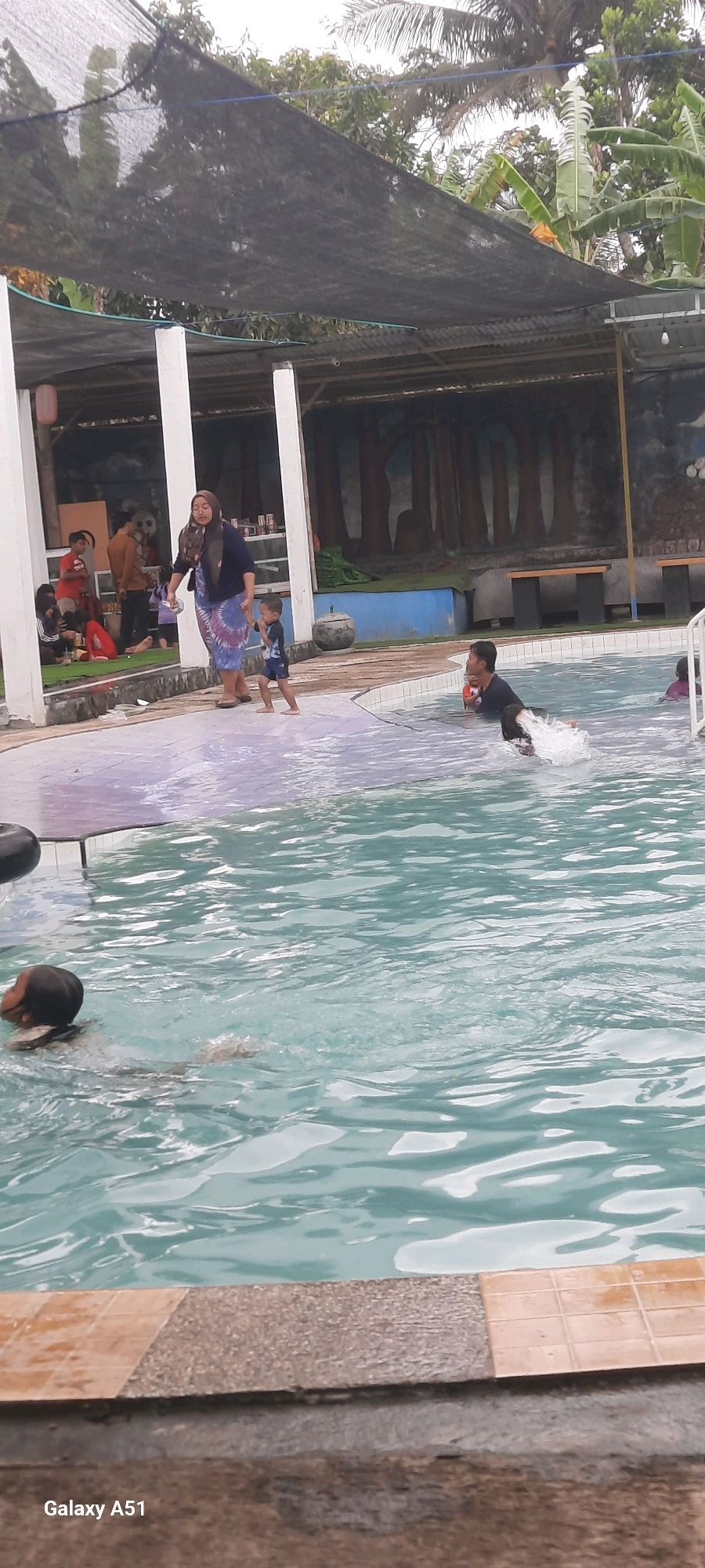 In Popoh, Indonesia, this image captures a portion of a swimming pool area. The foreground is dominated by the rippling surface of the turquoise pool water, with a light-colored tiled edge and a rougher, gray textured edge. In the middle ground, a woman in a headscarf and a patterned skirt is holding the hand of a young child wearing a blue swimsuit, walking near the pool's edge. To the right, a person is partially submerged in the water, creating a splash, and another child is visible further in the pool. The background reveals a shaded area with white pillars, a black netting overhead for shade, and what appears to be a mural or painted scene on a wall depicting trees. Several other individuals are seated or standing in the shaded area, suggesting a public or family gathering. The overall impression is a leisure scene at a water park or resort.