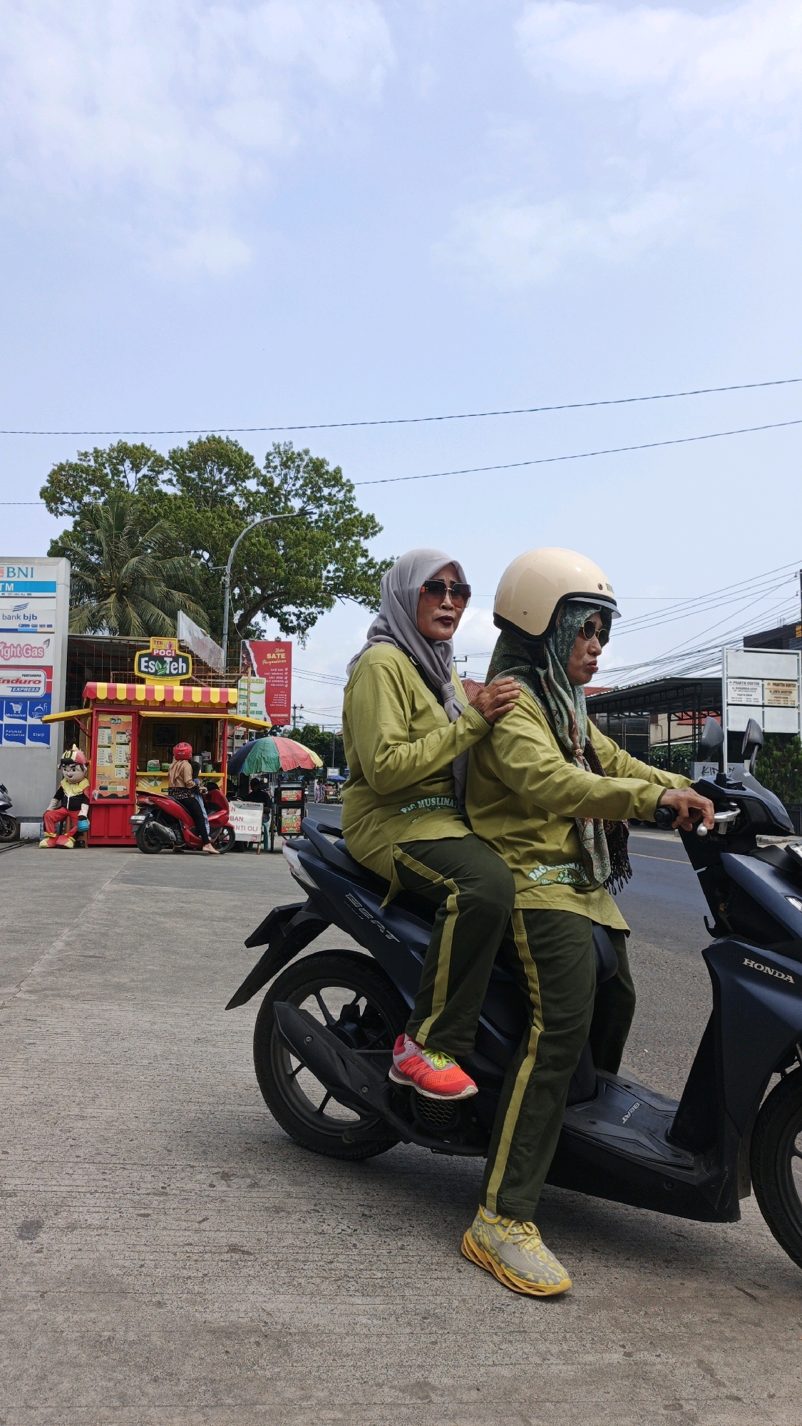 Two Indonesian women, wearing olive green long-sleeved shirts, dark green pants, and hijabs, are riding on a dark-colored scooter on a paved road in Pangandaran, Indonesia. The woman in front is driving and wearing a light beige helmet and sunglasses. The woman behind her is a passenger, wearing sunglasses and resting her left arm on the driver's shoulder. Both women are wearing athletic shoes. In the background, there is a red and yellow food stall with a sign that reads "Teh Poci Es Teh" and displays pictures of drinks. A statue of a man in a red and yellow uniform stands next to the stall. There are other motorcycles and a red scooter parked near the food stall. A large green tree is visible behind the food stall, and buildings and a blue sky with clouds are in the distance.