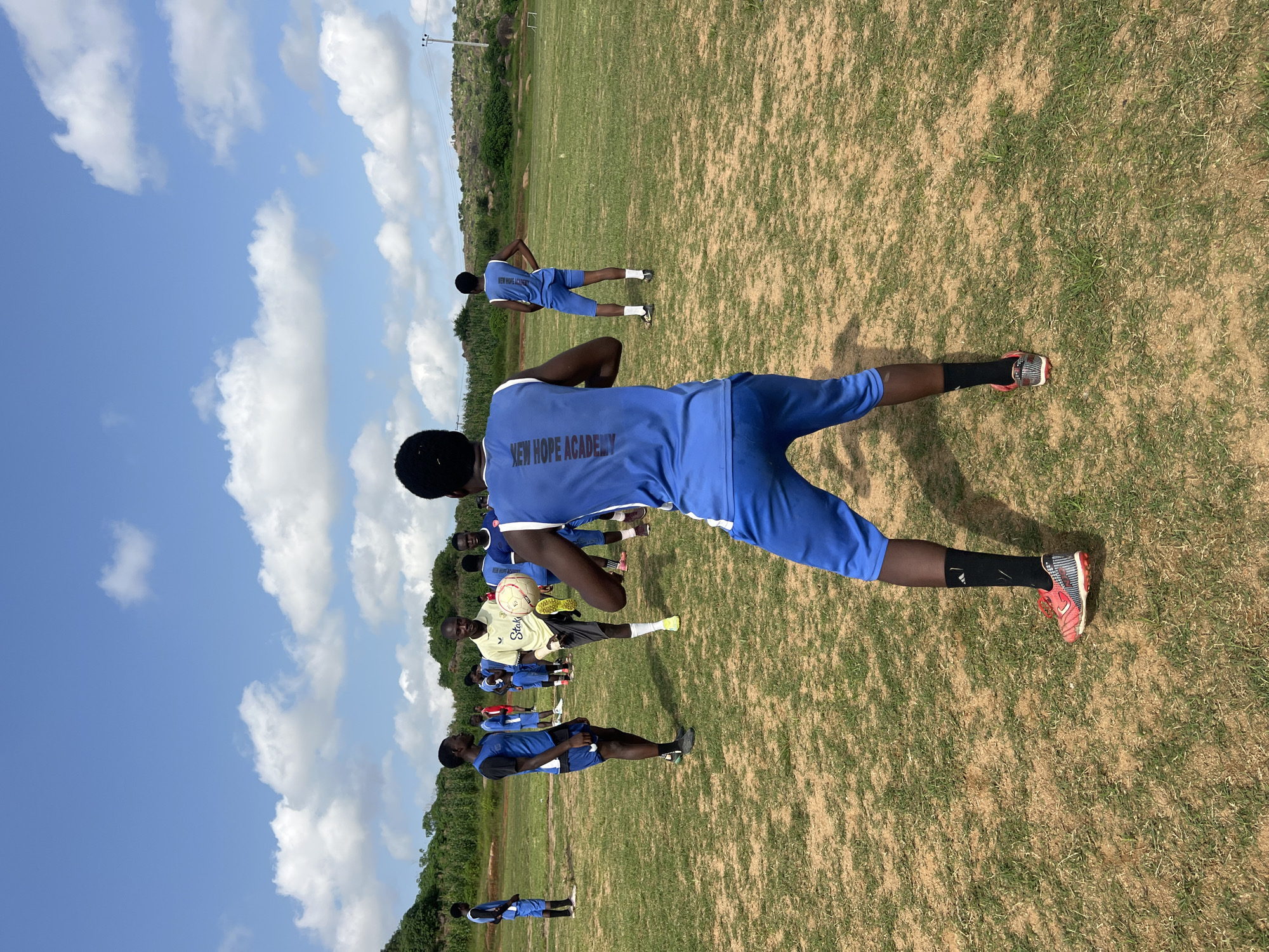 A group of young males in blue athletic uniforms are gathered on a grassy field under a partly cloudy sky. The setting appears to be an outdoor sports training ground, possibly in or near Jos, Nigeria, given the vegetation and terrain. In the foreground, a young male is positioned with his back to the camera, facing a group of his peers. He is wearing a blue sleeveless jersey with the text "NEW HOPE ACADEMY" in white lettering on the back, and blue shorts. His right leg is bent at the knee, and his right foot, clad in a red and white athletic shoe and a black sock, is extended towards a soccer ball.
To his left, another young male in a blue jersey and shorts stands with his hands on his hips, observing. Further back, a coach or instructor, wearing a yellow polo shirt and dark shorts, is kicking a soccer ball with his left foot. Several other young males in similar blue uniforms are scattered across the field, some appearing to be participating in drills or observing. The field itself is a mix of dry, light brown earth and sparse green grass. In the background, a gentle slope is covered with dry grass and scattered rocks, with some trees and vegetation visible further up. A utility pole with wires is also present in the distance. The overall scene depicts a football training session in an outdoor, somewhat arid environment.
