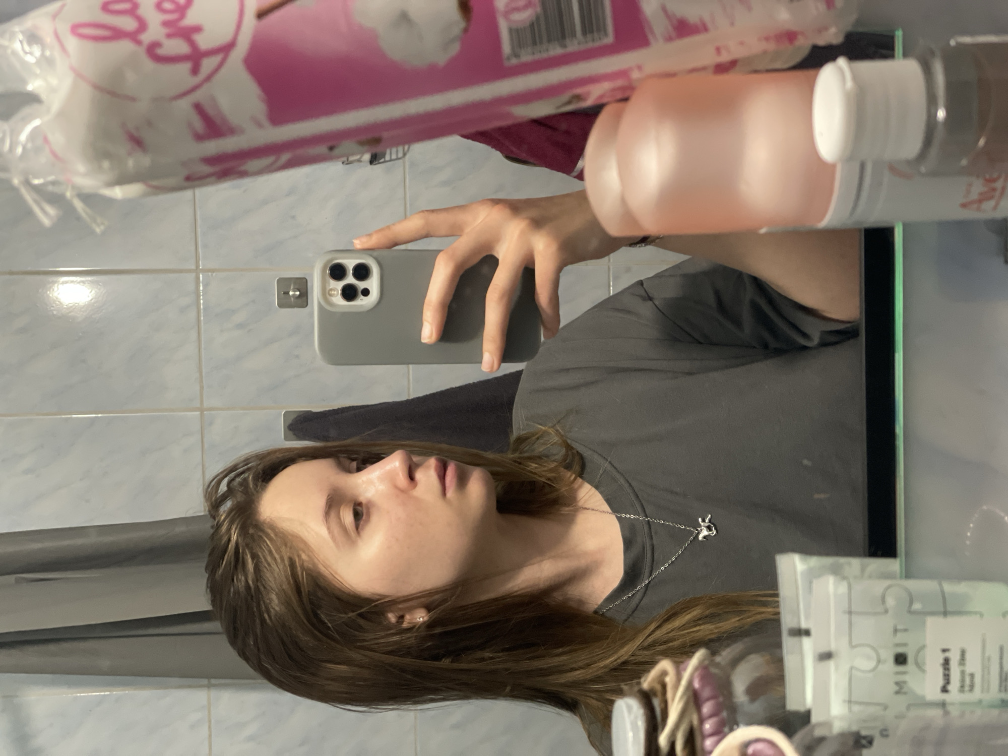 A young woman with brown hair is taking a mirror selfie in a bathroom. She is wearing a gray t-shirt and a silver necklace with a horse pendant. In her right hand, she is holding a gray iPhone. In the reflection of the mirror, there are several cosmetic products, including cotton pads and bottles of lotion. The bathroom walls are tiled with light blue tiles. The scene is indoors, likely in a residential setting in Chaikovsky, Russia.
