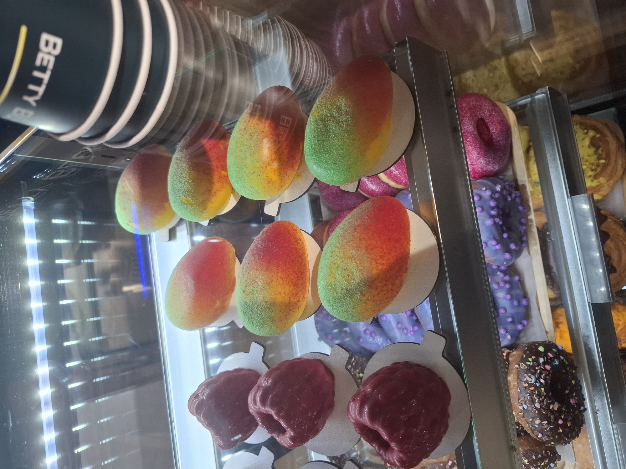 The image displays a close-up view of a refrigerated display case filled with pastries. The case is illuminated from above by white fluorescent lights, creating reflections on the glass. On the top shelf, there are several items that resemble mangoes, with a gradient of green, yellow, and red colors, and a textured coating. To the right, a stack of black paper cups with white rims is visible, with the "BETTY B" logo partially discernible on the side of the topmost cup. Below these mango-like pastries, on the second shelf, are red, raspberry-shaped pastries. Further down, on the third shelf, are donuts with purple icing and purple sprinkles, and a bright pink donut. The bottom shelf contains chocolate donuts with multi-colored sprinkles, and other pastries with yellow and green toppings. The overall environment appears to be a bakery or cafe, possibly in Dubăsarii Vechi, Moldova, given the mention of the city. There are no people visible in the image.