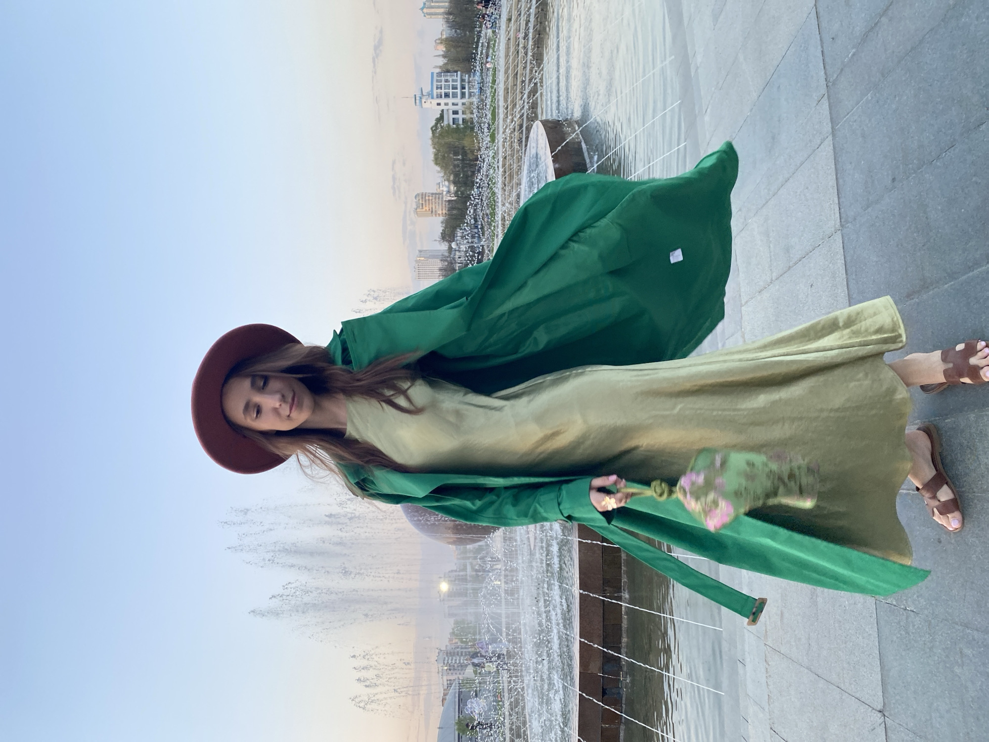 A woman stands next to a fountain. She wears a wide-brimmed brown hat, a light green dress, and a bright green trench coat. She holds a small green purse with pink floral detailing in her left hand. Her eyes are closed and she looks down. She is wearing brown sandals.
The fountain has numerous water jets spraying upwards. In the background, the city of Yekaterinburg, Russia, is visible, showing buildings and trees. The sky is light blue with a touch of sunset hue. The ground appears to be paved with gray stone slabs.