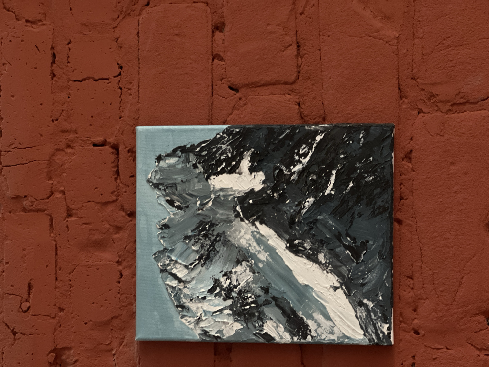 A landscape painting hangs on a red brick wall. The painting depicts a snowy mountain scene with heavy impasto texture. The background of the painting is light blue, and the mountain itself is rendered in shades of white, gray, and black. The brick wall has visible texture and imperfections. The location is unknown.