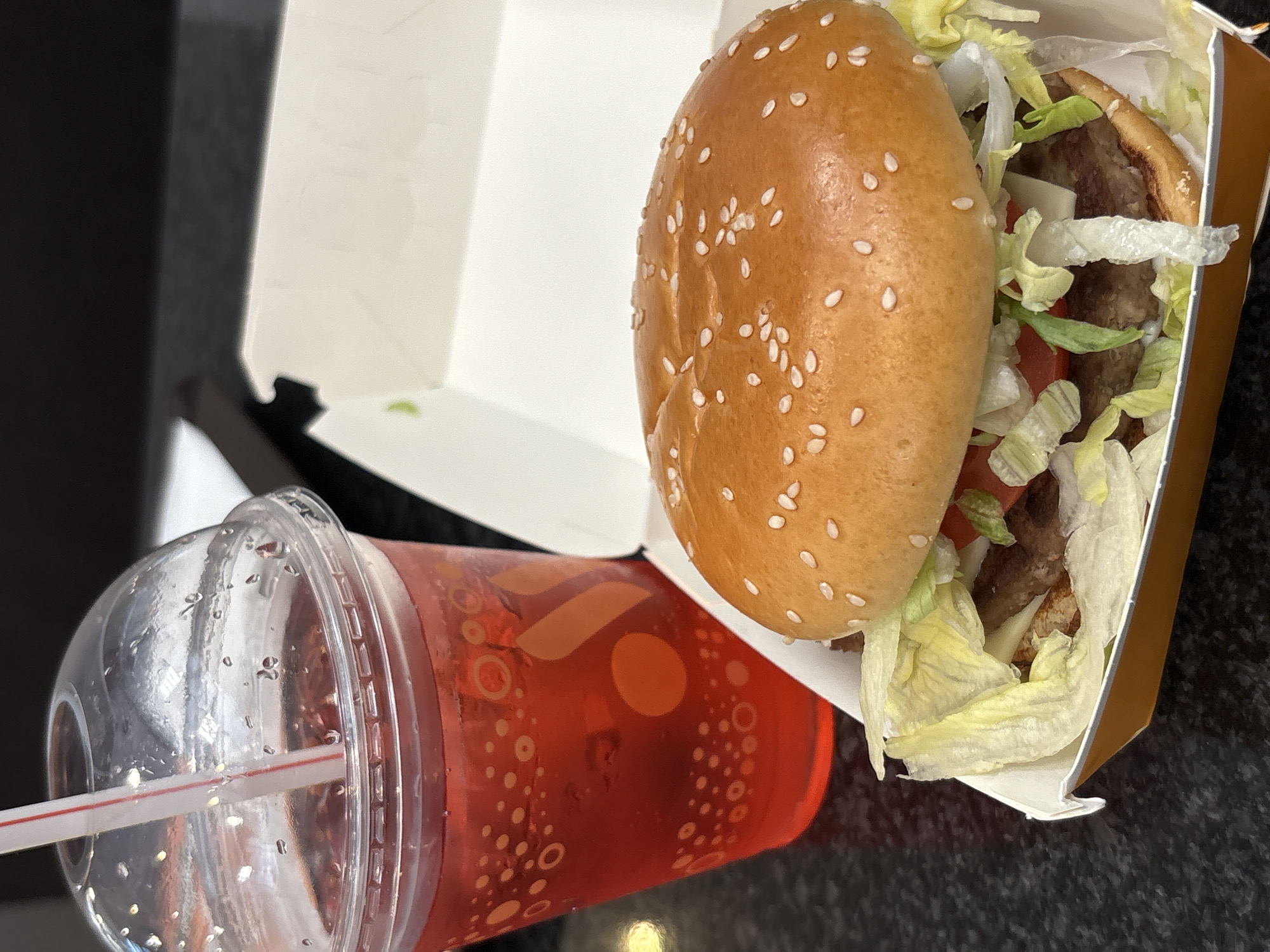 A close-up, eye-level shot shows a hamburger and a red beverage with a straw, placed on a black speckled surface. The hamburger is served in a white and orange cardboard box. The burger bun is sesame-seeded and golden brown, with visible lettuce, tomato slices, and possibly onions. The red beverage is in a clear plastic cup with condensation on the outside. The cup has a white and red striped straw inserted into it and features a pattern of orange circles and dots. The background is dark and out of focus. The scene appears to be indoors, possibly at a fast-food establishment. The location is identified as Saint Petersburg, Russia.