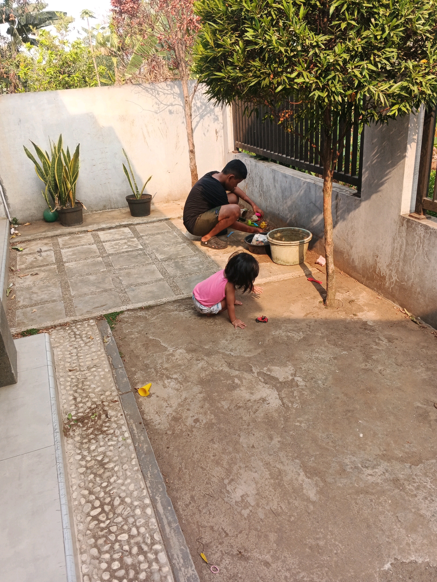 The image depicts an outdoor scene in Kadu, Indonesia, likely a residential backyard. A man, dressed in a black t-shirt and shorts, is kneeling on the ground to the right, engaged with some objects in a dark basin. Beside him, a young child in a pink shirt and shorts is crawling on the dirt, focused on a small red toy car. Behind the man, a large tree with green foliage provides shade, and a metal fence is visible. To the left, a concrete wall with two potted snake plants stands against a backdrop of lush green vegetation. The ground is a mix of concrete tiles and packed dirt. The lighting suggests it is daytime, with sunlight casting shadows. A yellow funnel-shaped object and some scattered leaves are on the ground in the foreground.