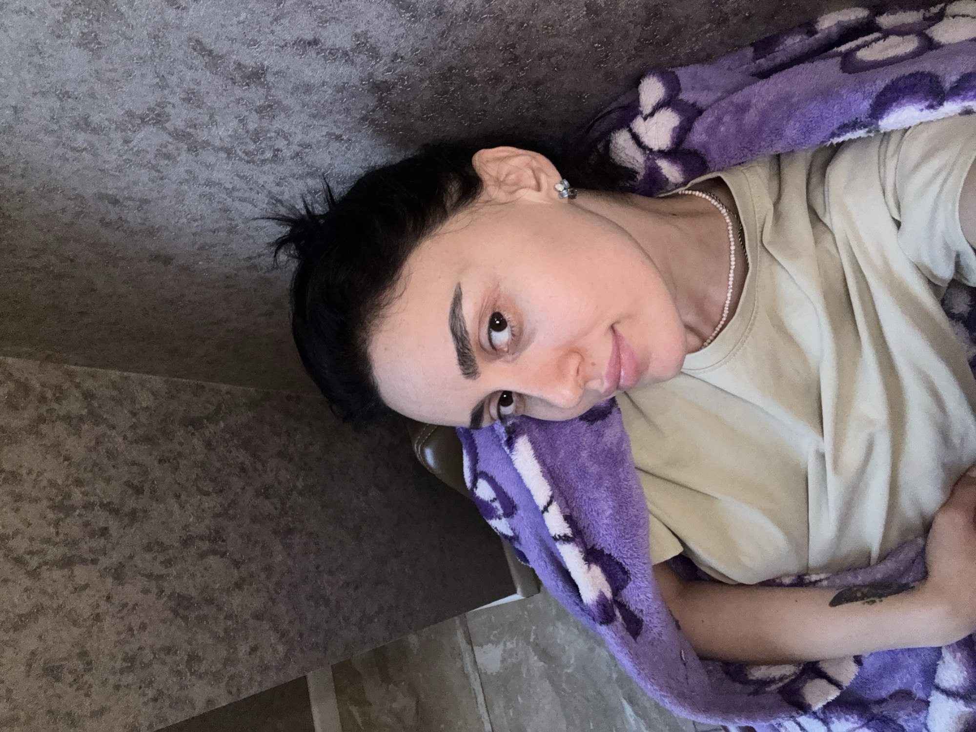 The image displays an adult female in an indoor setting, looking directly at the camera. She has dark hair, dark eyebrows, and is wearing a light beige t-shirt. A delicate light-colored beaded necklace is visible around her neck, and a small stud earring is present in her right ear. Her left forearm features a dark, vertical abstract tattoo. She is partially covered by a purple plush blanket with a repeating lighter-colored floral or leaf pattern. The background is a textured wall with mottled grey and dark brown tones. A segment of dark brown furniture and a light-colored baseboard are visible in the lower left background. The specific location within Tbilisi, Georgia, cannot be determined from the visual evidence.