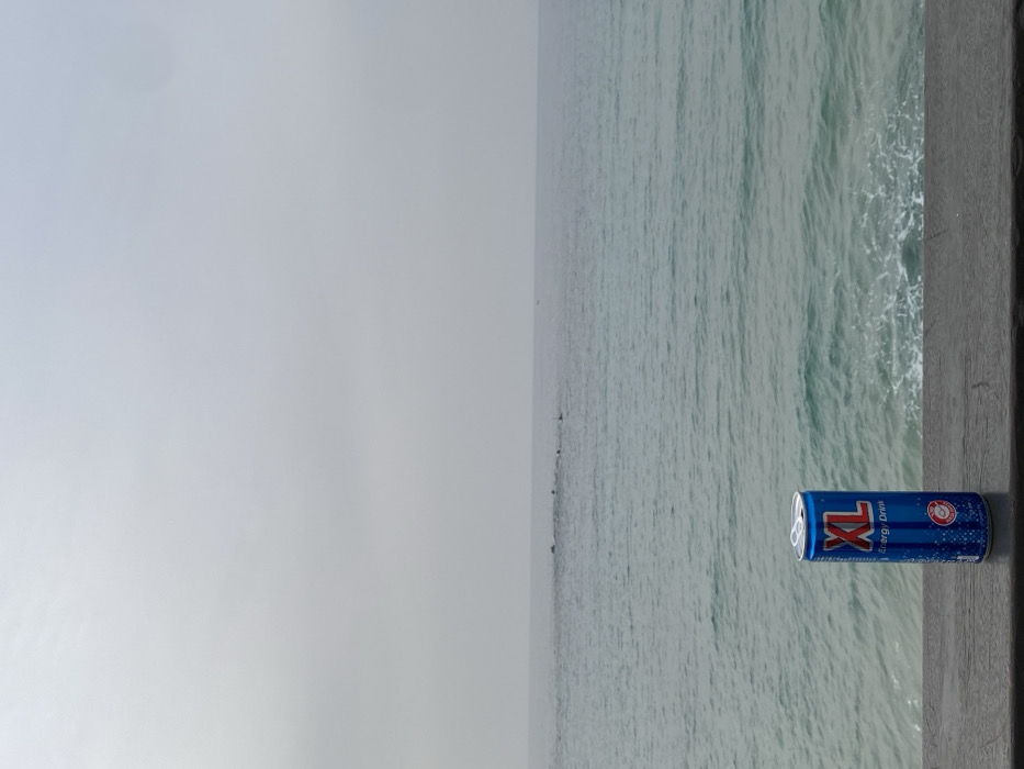 A blue XL Energy Drink can stands upright on a dark, weathered wooden railing or surface in the foreground, centered horizontally. The can features prominent red and white "XL" branding with "Energy Drink" written below. Beyond the railing, the Mediterranean Sea extends under an overcast sky in Tel-Aviv, Israel. The sea water is a muted blue-green with gentle ripples and minor whitecaps near the foreground. In the mid-distance, a line of low-lying, dark structures, consistent with a breakwater or submerged rocks, is visible stretching horizontally across the water, accompanied by one or two small, dark buoys. The sky is uniformly light grey, indicating an overcast condition, meeting the sea at a subtle horizon line. No individuals, animals, or active subjects are present in the frame.