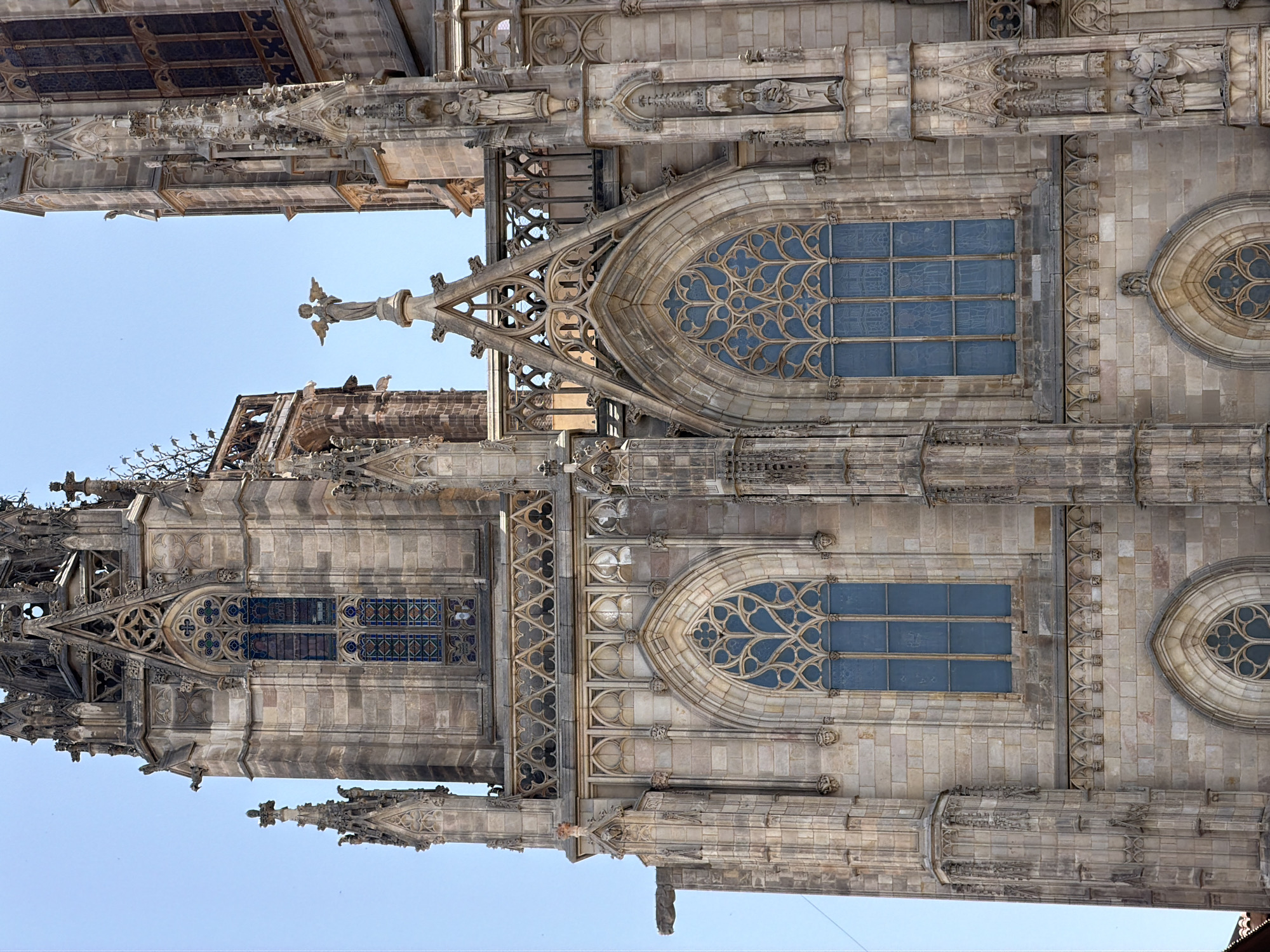 The image shows a close-up, eye-level view of the facade of the Barcelona Cathedral in Barcelona, Spain. The cathedral is a Gothic architectural masterpiece, constructed primarily from stone. The scene is dominated by intricate stone carvings, pointed arches, and stained-glass windows.
On the left, a tall, slender spire with decorative pinnacles and finials rises against a clear blue sky. Gargoyles protrude from the sides of the building, characteristic of Gothic architecture. Below these spires, large pointed arch windows with complex tracery are visible, filled with blue stained glass.
In the center of the image, a statue of an angel with outstretched wings stands atop a spire, reaching towards the sky. This angelic figure is a prominent detail on the cathedral's facade.
To the right of the angel, another section of the cathedral rises, featuring more pointed arch windows and elaborate stone ornamentation. Statues of figures, likely saints, are integrated into niches along the facade.
The stonework appears weathered, indicating the age of the structure. The lighting suggests a bright, sunny day, casting subtle shadows on the detailed carvings and enhancing the texture of the stone. The overall impression is one of grandeur and intricate craftsmanship, typical of a historic European cathedral.