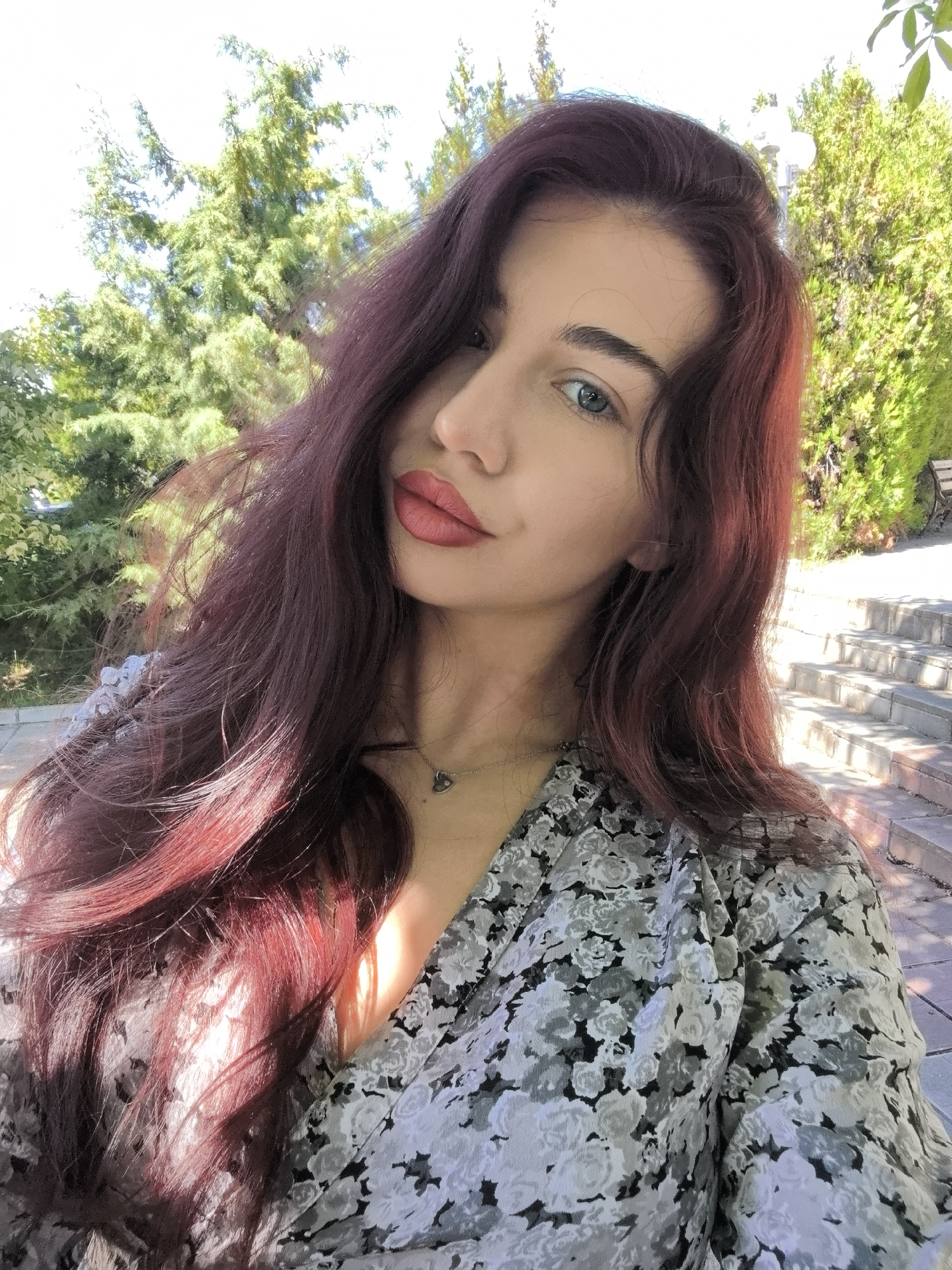 A young woman with long, burgundy-colored hair, light eyes, and red lipstick is the subject of the image. She is wearing a gray and white floral-patterned dress. A delicate necklace is visible around her neck. The background consists of greenery, with trees visible behind her and a stone staircase on the right side of the frame. The location appears to be an outdoor area, possibly a park or garden. The city is unknown.