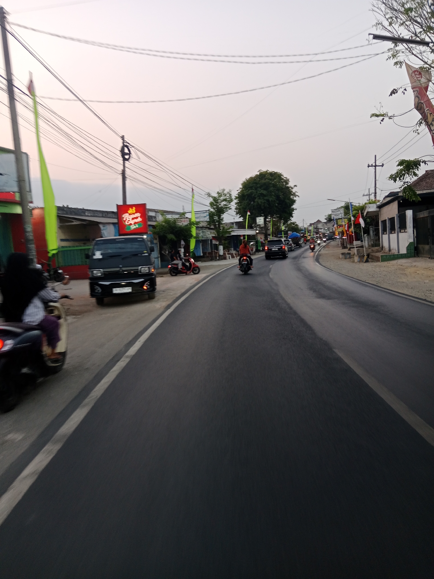 The image depicts a street scene in Ringinrejo, Indonesia, at what appears to be dusk. The road is paved and has a white dividing line. Several vehicles are present, including a black van, a black car, and multiple motorcycles. One motorcycle in the foreground is partially visible, with a rider wearing a black headscarf and white and purple clothing. Another motorcycle further down the road has a rider in an orange shirt. A red scooter is parked on the side of the road. Buildings line the street, some with signs, including a red sign with yellow lettering. Tall poles with numerous wires are scattered throughout the scene, indicating overhead electrical infrastructure. Green and red flags are visible, attached to poles and some buildings. A large tree is situated on the left side of the road, and other foliage is present. The sky is a light, hazy color, suggesting either overcast conditions or the end of the day. The overall impression is a typical roadside environment in a populated area of Indonesia.