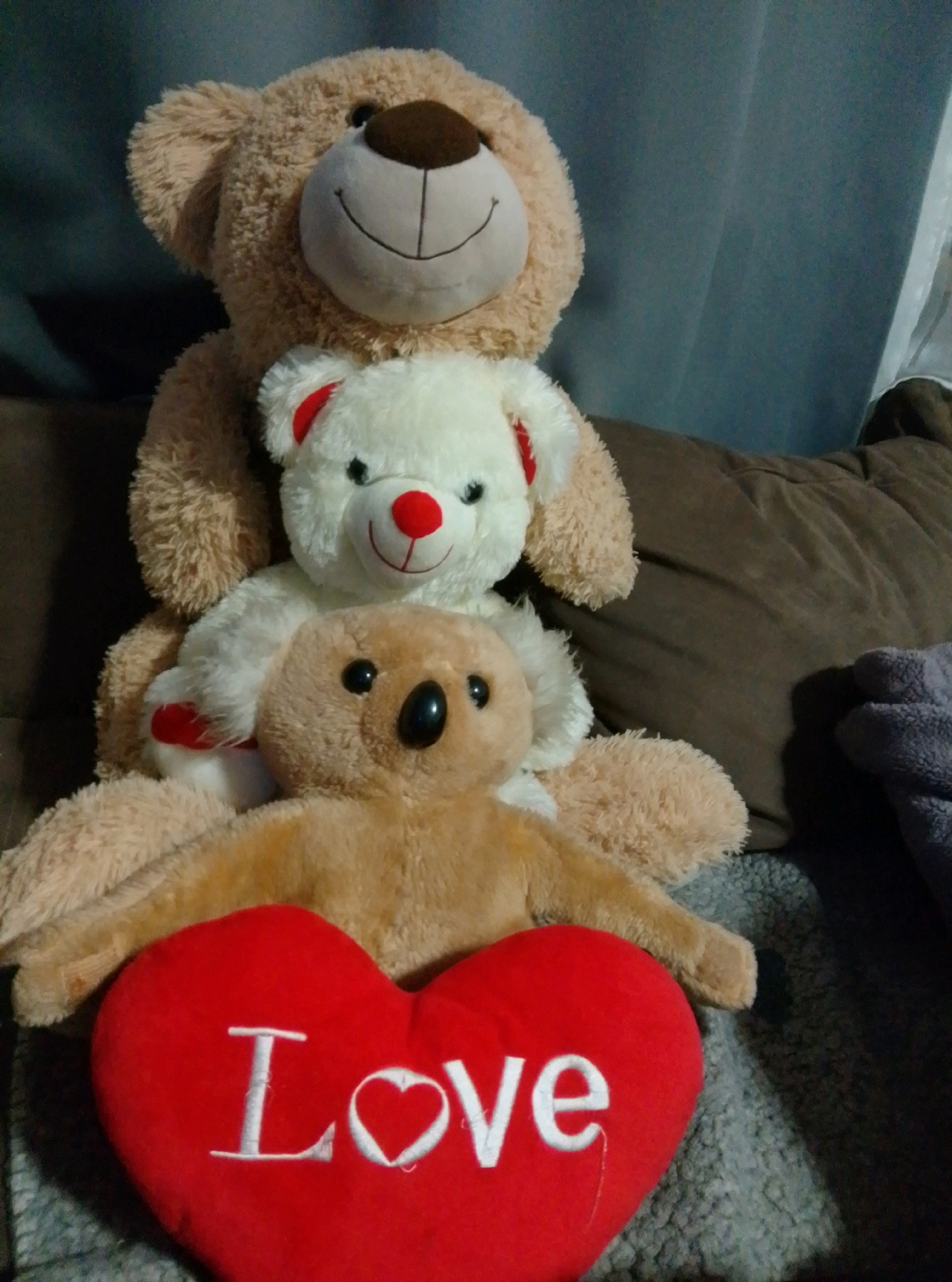 Three plush teddy bears are stacked vertically on a couch. The largest bear, a light brown one, is at the top, facing forward with a smiling expression. Below it is a smaller white bear with red ears and a red nose, also facing forward. The bottom bear is a light brown koala bear. In front of the koala bear is a red heart-shaped pillow with the word "Love" embroidered in white. The background consists of a dark gray couch and a gray textured surface underneath the bears. There is no discernible location in Wedel, Germany.