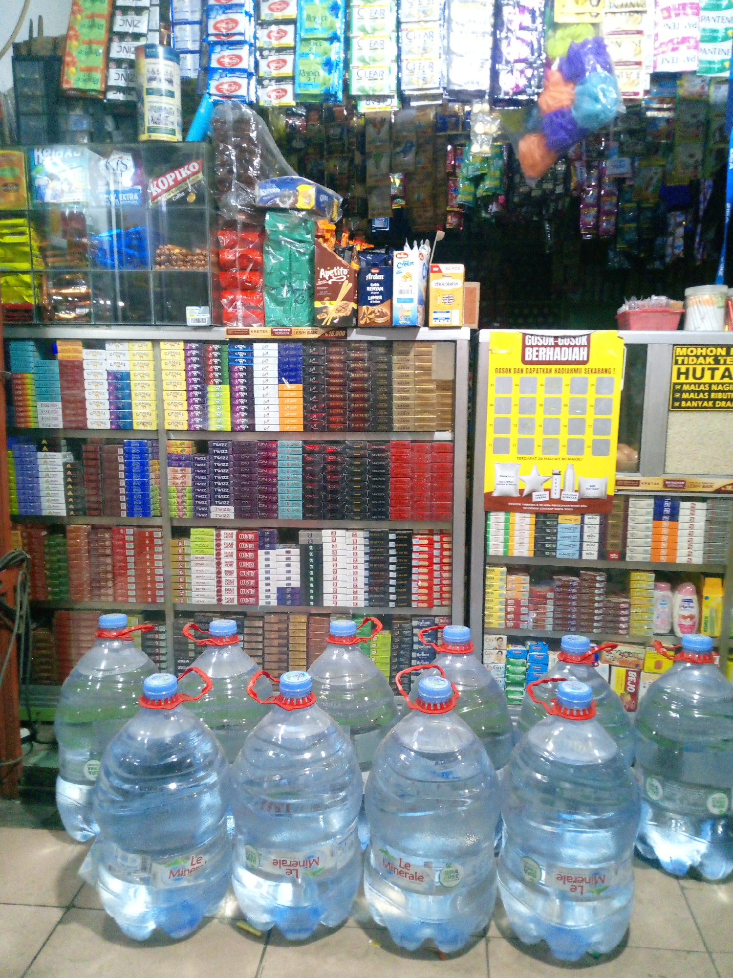 This is a medium shot, eye-level, indoor photograph of a small convenience store, likely a warung, located in Bitung Jaya, Indonesia. The store is densely packed with merchandise, predominantly food and drink items, displayed on shelves.
In the foreground, five large, clear plastic gallon jugs of "Le Minerale" bottled water are arranged on the tiled floor. The jugs have red plastic caps and blue dispenser spouts. The label "Le Minerale" is visible on each jug.
Behind the water jugs, numerous shelves are filled with various products. On the left side, there are rows of cigarette packs, including brands like "Camel," "Esse," and "LA." Further up are various snack packets and packaged goods. In the center, a large yellow promotional sign with the text "GUSUK-GUSUK BERHADIAH" (Scratch-Scratch for Prizes) is prominently displayed. Below this sign are shelves stocked with more cigarette packs in different colors and brands such as "Twizz" and "ON." On the right side, there are more shelves filled with packaged goods, including soap or shampoo bottles and boxes of rice. A small red basket with white cotton swabs is also visible.
The overall environment appears to be a typical small retail space in Indonesia, characterized by its extensive product variety and organized shelving. The lighting is adequate, illuminating the merchandise. No people are visible in the frame.