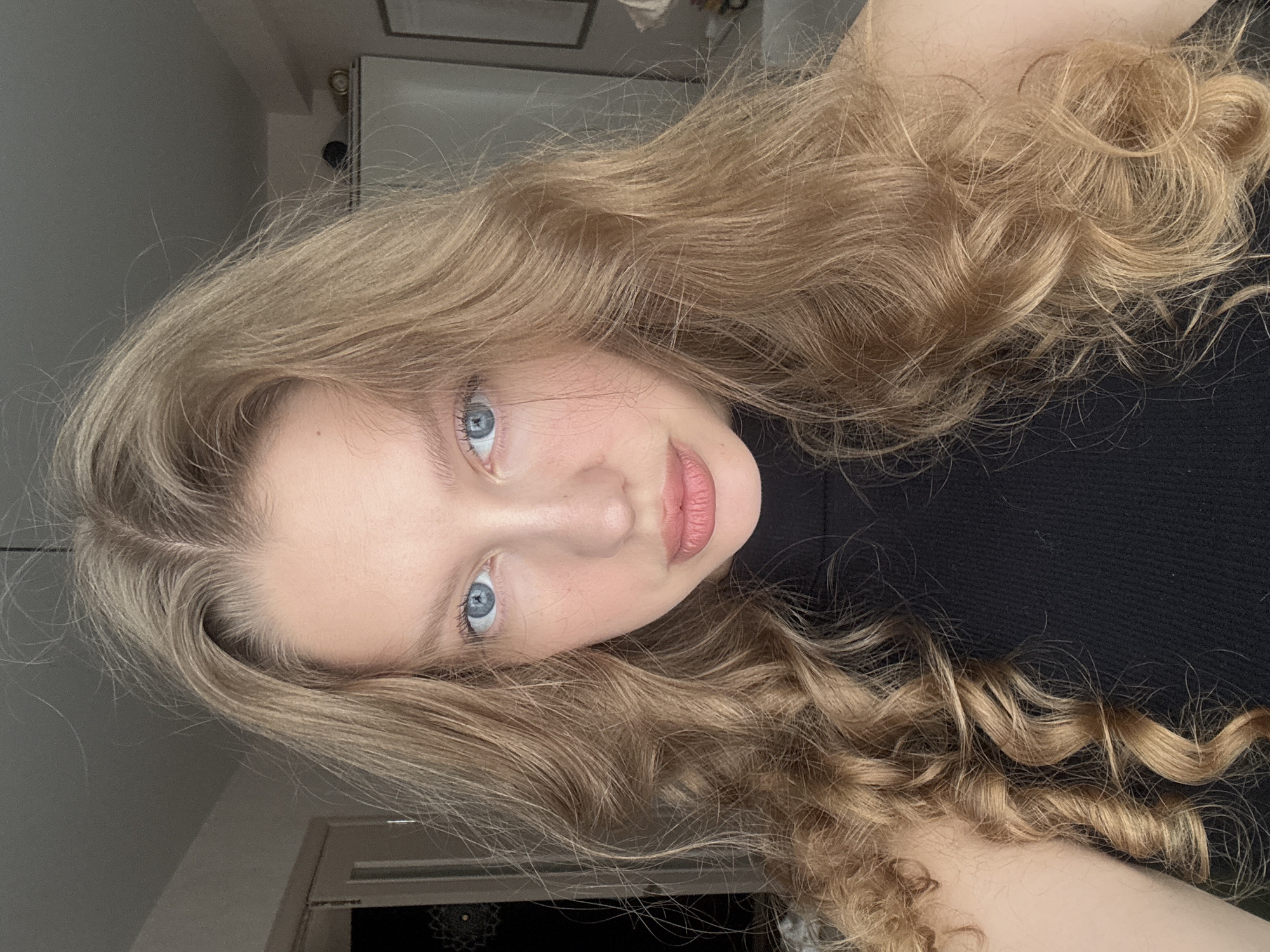 The image is a headshot of a young woman with blonde, wavy hair. She is looking directly at the camera, with her head slightly tilted. Her eyes are blue, and she has light skin. She is wearing a black turtleneck top. The background is an indoor setting, possibly a bedroom or living room, with muted colors and soft lighting. There are hints of furniture and a door in the background, suggesting a residential interior. No specific location in Izhevsk, Russia can be identified from the image.