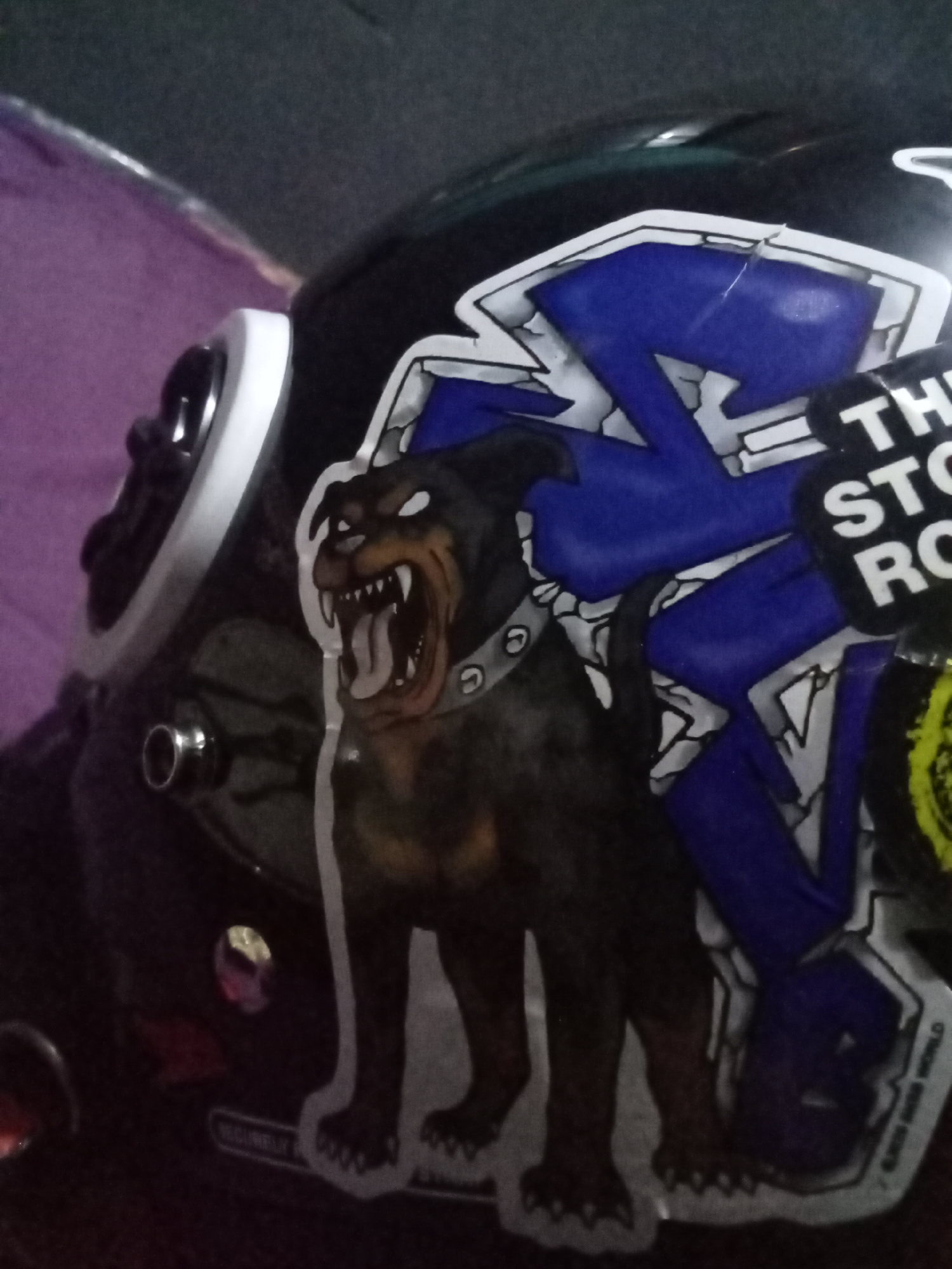 A close-up, slightly blurry, eye-level shot shows a black helmet with various stickers on its surface. The helmet is positioned on a purple, textured surface.
Dominating the frame is a large sticker depicting a fierce-looking Rottweiler with its mouth open, sharp teeth bared, and its tongue hanging out. The dog is primarily brown and black, with a gray spiked collar. To the right of the dog sticker is a graphic sticker with bold blue angular shapes on a white background, outlined in black. Part of a black sticker with white text is visible, with "TH", "STO", and "RO" being legible. There are also some faint yellow and green circular elements on the far right.
Visible on the helmet are two silver circular fasteners, likely for straps or ventilation. A white band encircles the front edge of the helmet. A portion of a small red decal is visible on the lower left side of the helmet, beneath the dog sticker. The background is dark and indistinct, with hints of purple fabric. The overall lighting is dim.