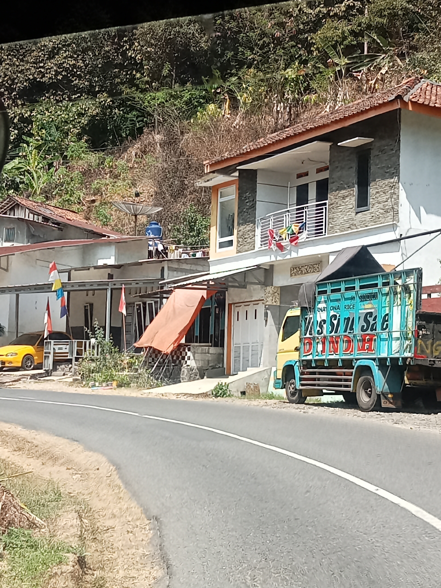 The image shows a street scene in Gambarsari, Indonesia. In the foreground, a paved road with a white line curves to the right. On the left side of the road, dry grass and sparse green vegetation are visible. In the middle ground, several buildings are situated along the road. To the left, a yellow car is parked in front of a low-rise building with a tiled roof. Several Indonesian flags are displayed on poles in front of this building. Further to the right, a two-story building with a tiled roof and gray stone facade is visible. In front of this building, a light blue truck with a turquoise cargo bed is parked. The truck has writing on its side, including "INTERNATIONAL RICE" and "DINDIN." A black tarp covers the top of the truck's cargo. Behind the buildings, a hillside covered in dense green vegetation rises steeply. A satellite dish is mounted on the roof of one of the buildings. The overall scene depicts a rural or semi-rural environment with a clear sky and sunlight illuminating the area.