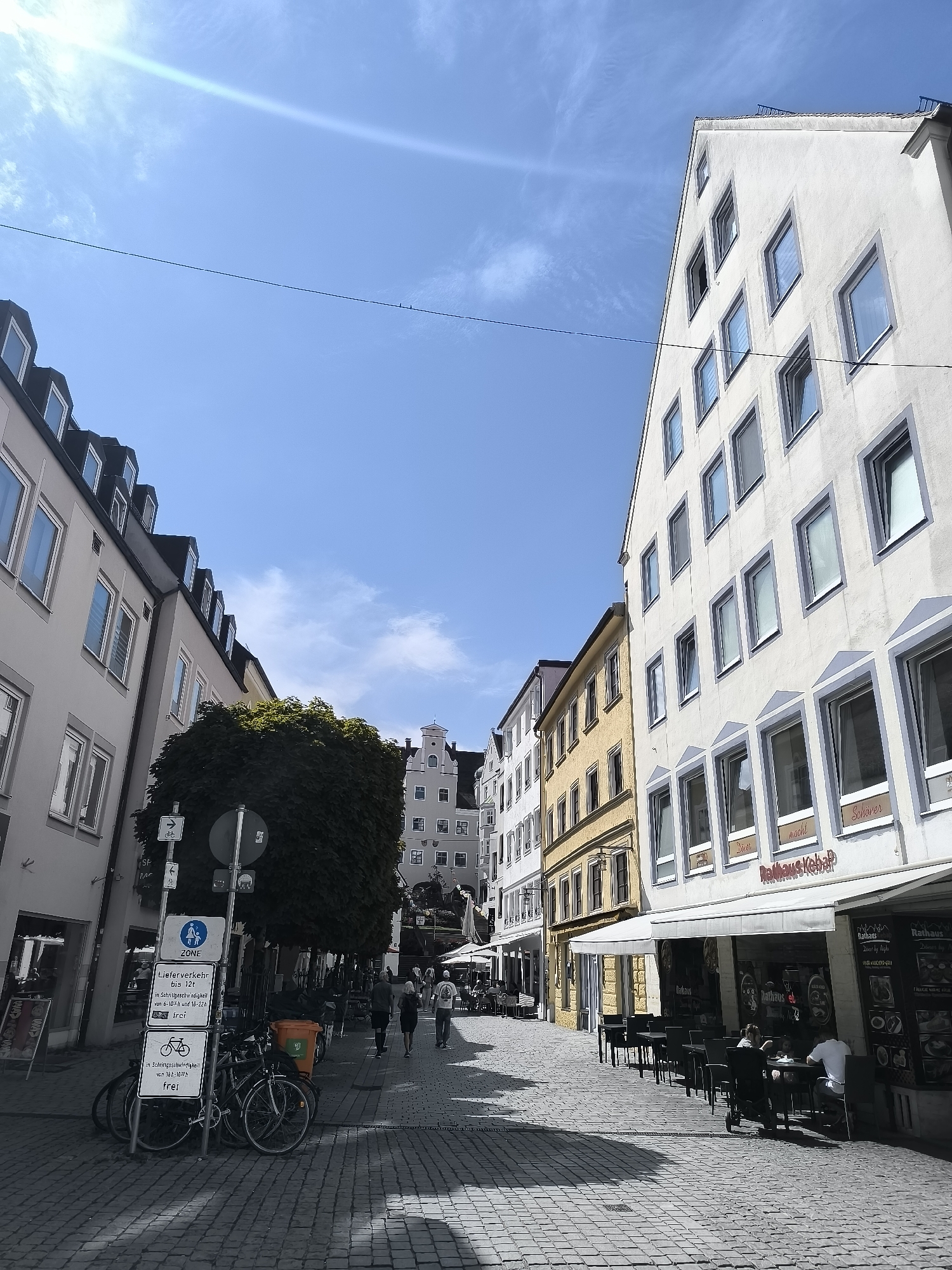 The image depicts a narrow cobblestone street in Kempten (Allgäu), Germany, lined with multi-story buildings. On the right, a white building features numerous windows with gray trim and signs for "Rathaus Kebab" and "Schöber". An awning extends over a row of outdoor tables and chairs, where two individuals are seated. Further down the street, more tables and chairs are visible, suggesting a bustling outdoor dining area. On the left, a light gray building with dormer windows is visible, followed by a yellow building. A large tree with dense foliage stands in the middle ground on the left. Several bicycles are parked near a signpost with traffic regulations. People are walking along the street, some in groups, and others individually. The sky is clear blue with some wispy clouds. The scene captures a typical urban streetscape with a mix of commercial establishments and pedestrian activity under bright sunlight.