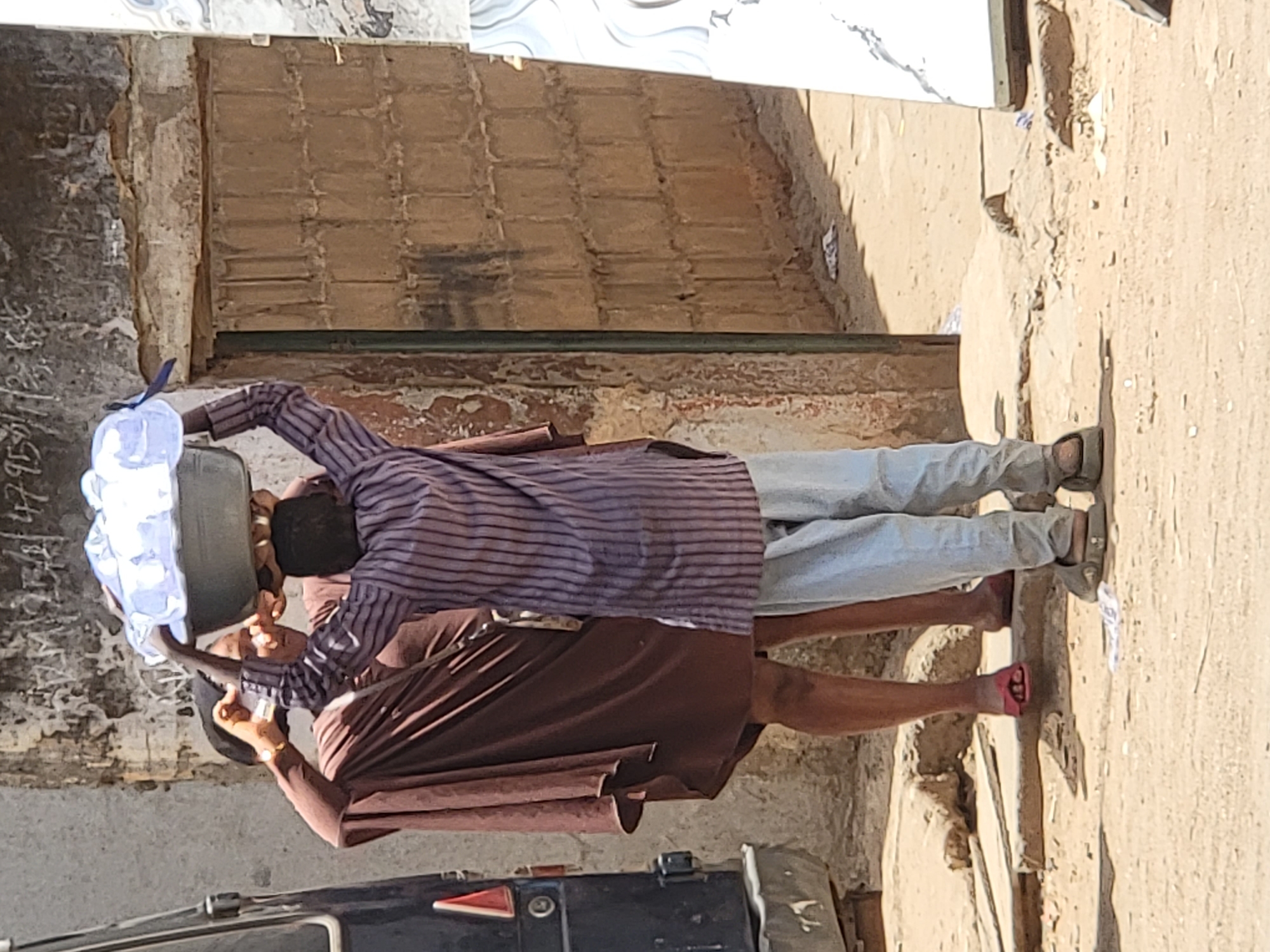 In Maiburiji, Nigeria, two individuals are carrying a large container together in an outdoor urban environment. One individual, wearing a striped shirt and light-colored jeans, holds one side of the container. The other individual wears a brown shirt. The container appears to be filled with plastic bags, possibly containing water or ice. They are standing on a concrete surface near a building. A vehicle is partially visible at the bottom of the image.