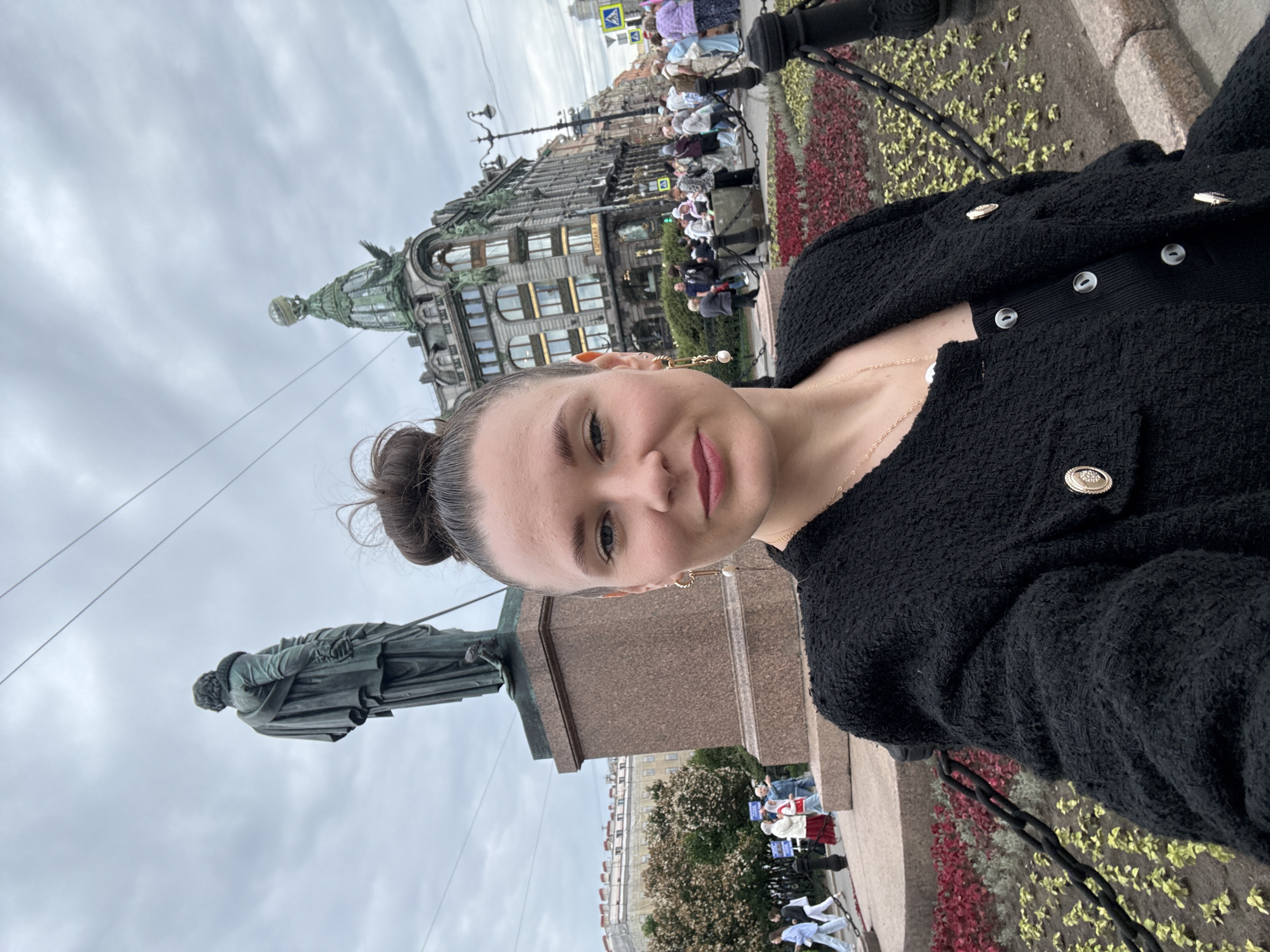 A woman takes a selfie in front of a monument and a historic building. The woman has her hair in a bun and is wearing a black jacket with gold buttons and a black top. She is also wearing earrings and a necklace. In the background, a bronze statue of a man stands on a granite pedestal. Behind the statue is a large, ornate building with a distinctive green dome and a globe on top. People are walking around in the background, suggesting a public space in a city. The scene appears to be in Saint Petersburg, Russia, likely near Nevsky Prospekt, as the building is the Singer House, a well-known landmark.