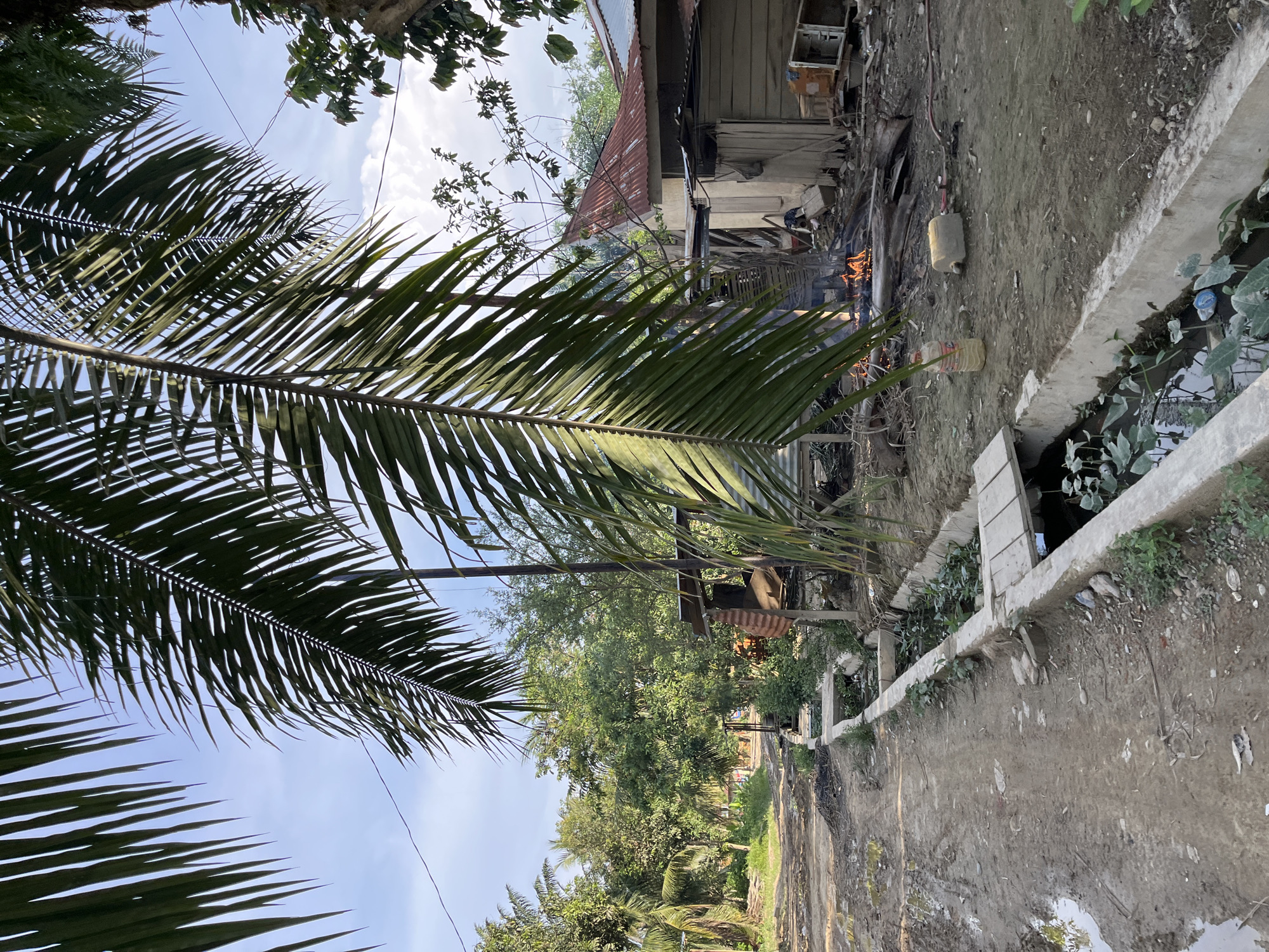 A scene in a rural area of Landuh, Indonesia, depicts a muddy, dirt road with puddles of water to the left. A concrete drainage ditch runs parallel to the road on the right, filled with murky water and aquatic plants. Beyond the ditch, a dilapidated structure with a rusted tin roof is visible. In the foreground right, a plastic bottle and a beige jerry can sit on the ground. A small fire burns in the middle ground, surrounded by debris and discarded items. Further in the background, more houses with tin roofs and lush green trees create a tropical ambiance. The sky is partly cloudy.